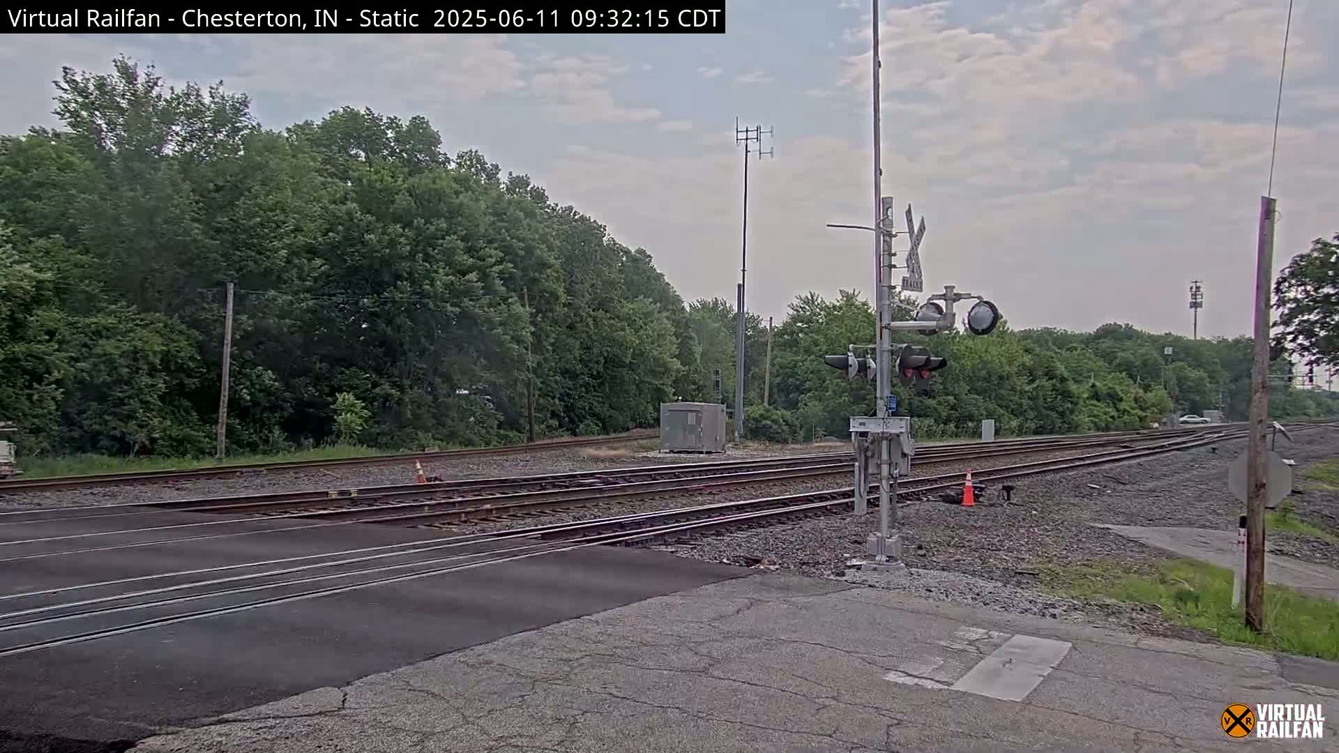 A railroad crossing with a signal light and several sets of tracks is surrounded by lush green trees under a partly cloudy sky.