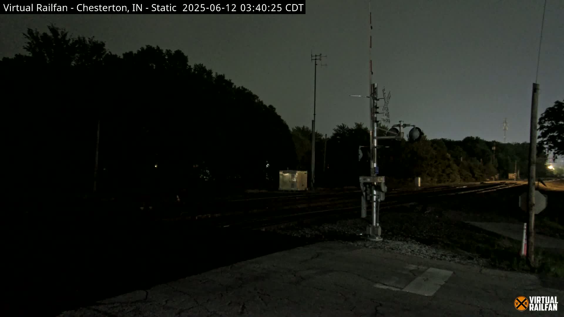 A nighttime view of a railroad crossing with train tracks extending into a dark, tree-lined area under a cloudy sky.