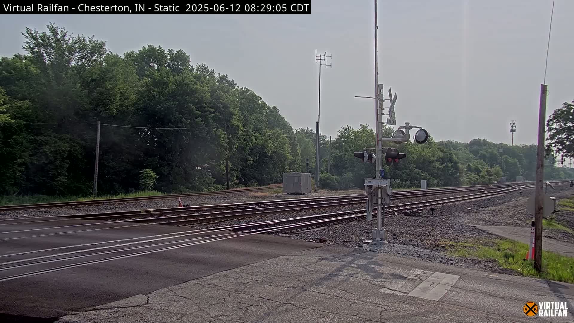 A railroad crossing with multiple tracks is seen on an overcast day, bordered by lush green trees.