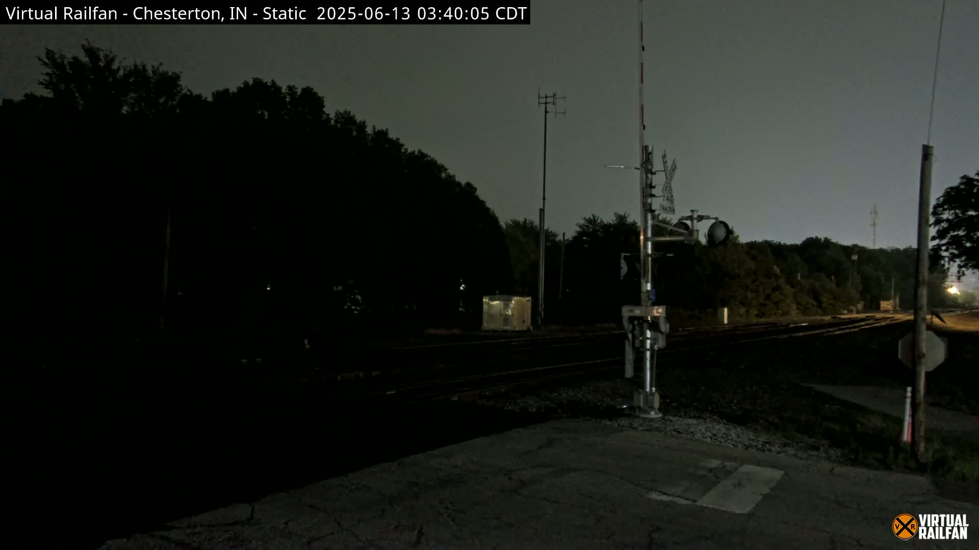 A nighttime view of a railroad crossing with dark trees in the background under a gray sky.
