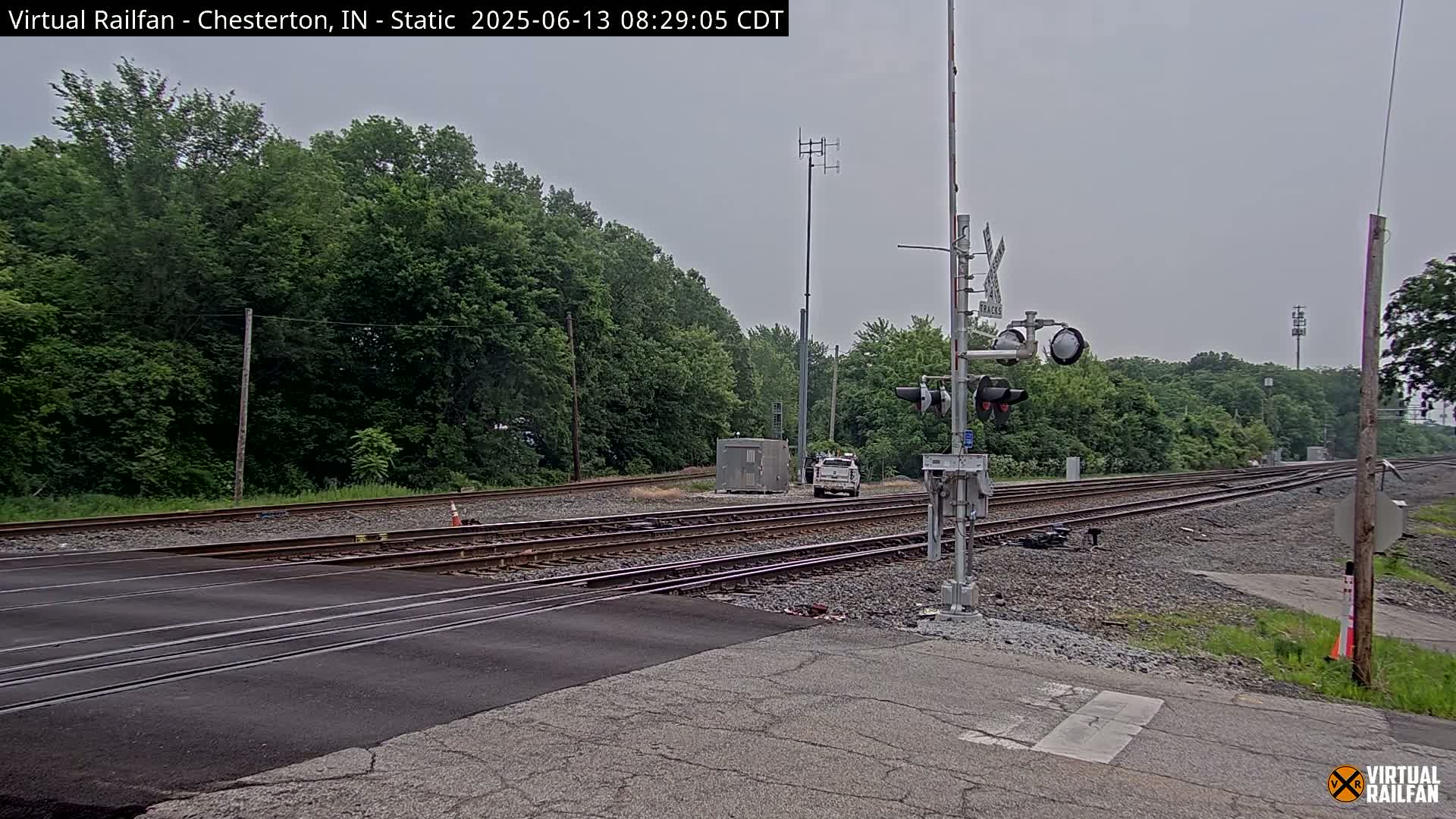 A railroad crossing with multiple tracks is shown, under an overcast sky, surrounded by lush green trees and some utility poles.