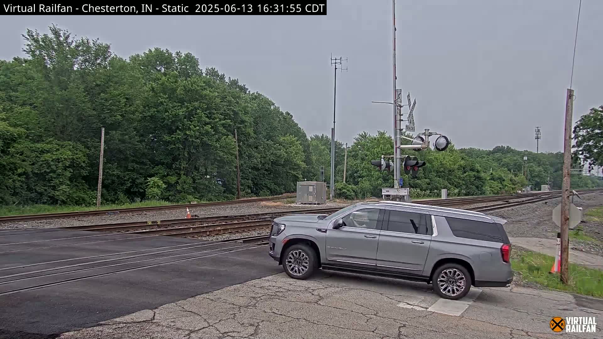 A silver SUV is stopped at a railroad crossing on an overcast day, with green trees lining the tracks in the background.