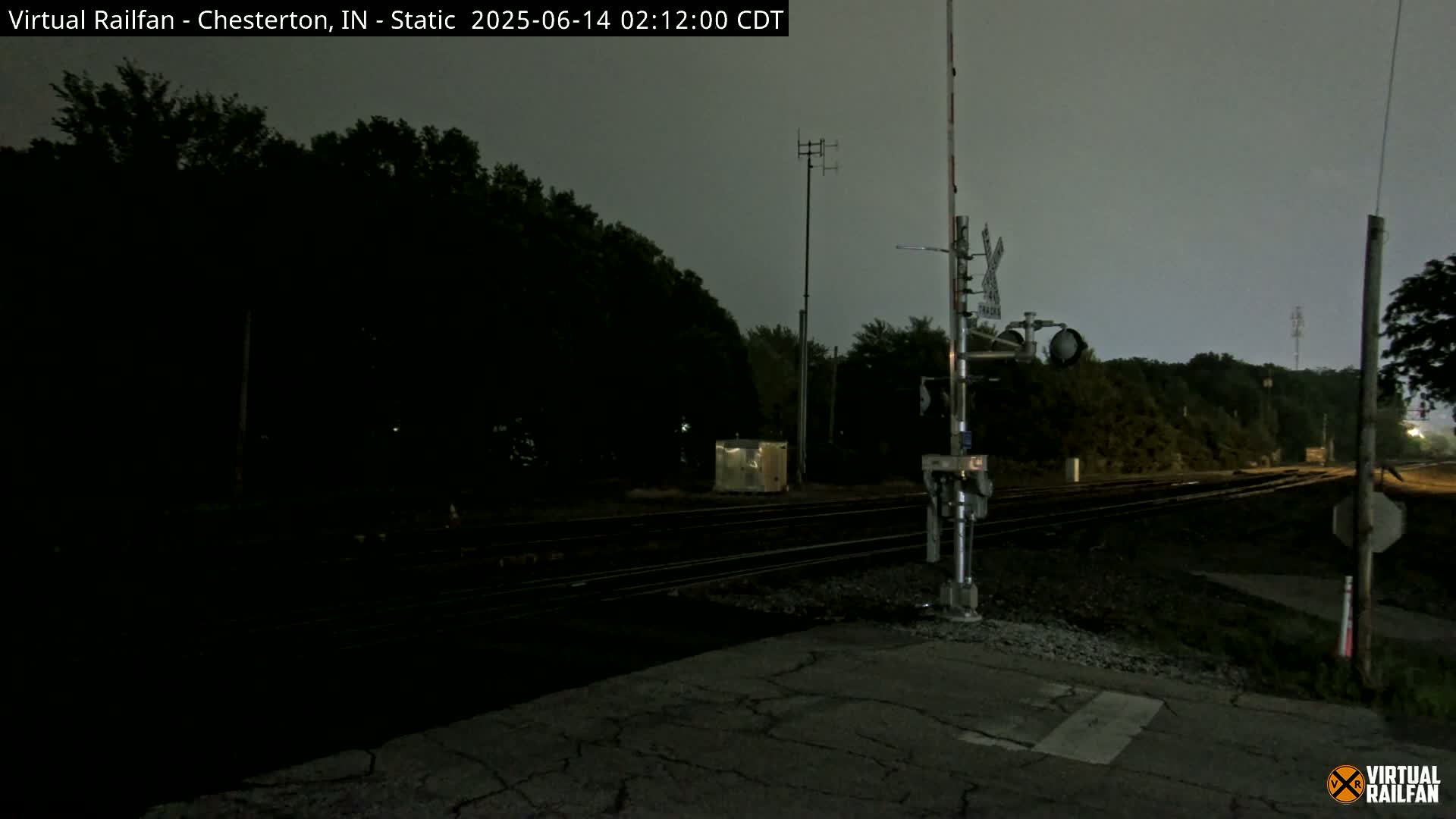 A dark, nighttime view of a railroad crossing under a gray sky shows train tracks, signals, and surrounding trees.