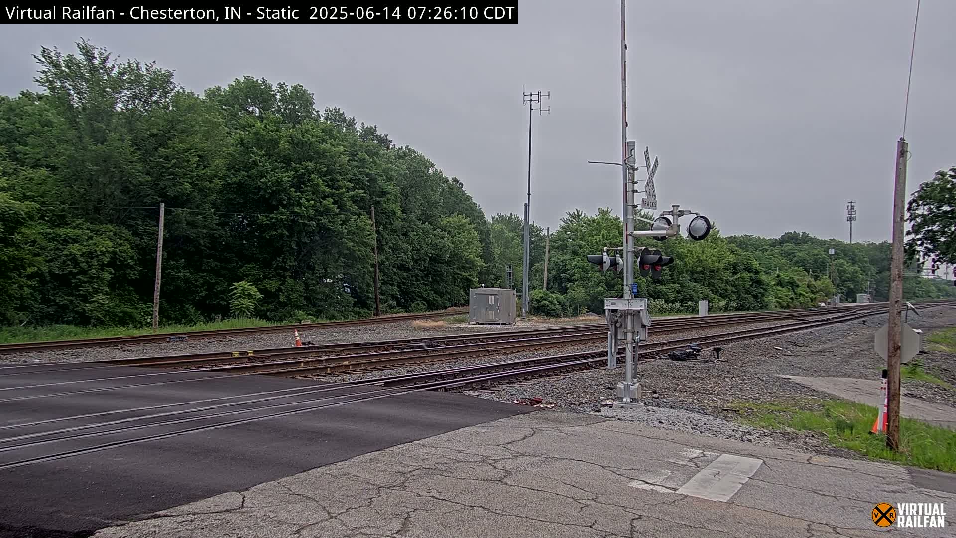 A railroad crossing with traffic signals and a backdrop of lush green trees under an overcast sky.
