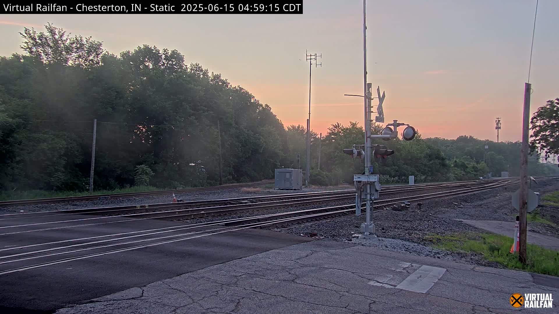 A railroad crossing with multiple tracks under a pinkish-orange sunrise, surrounded by lush green trees.