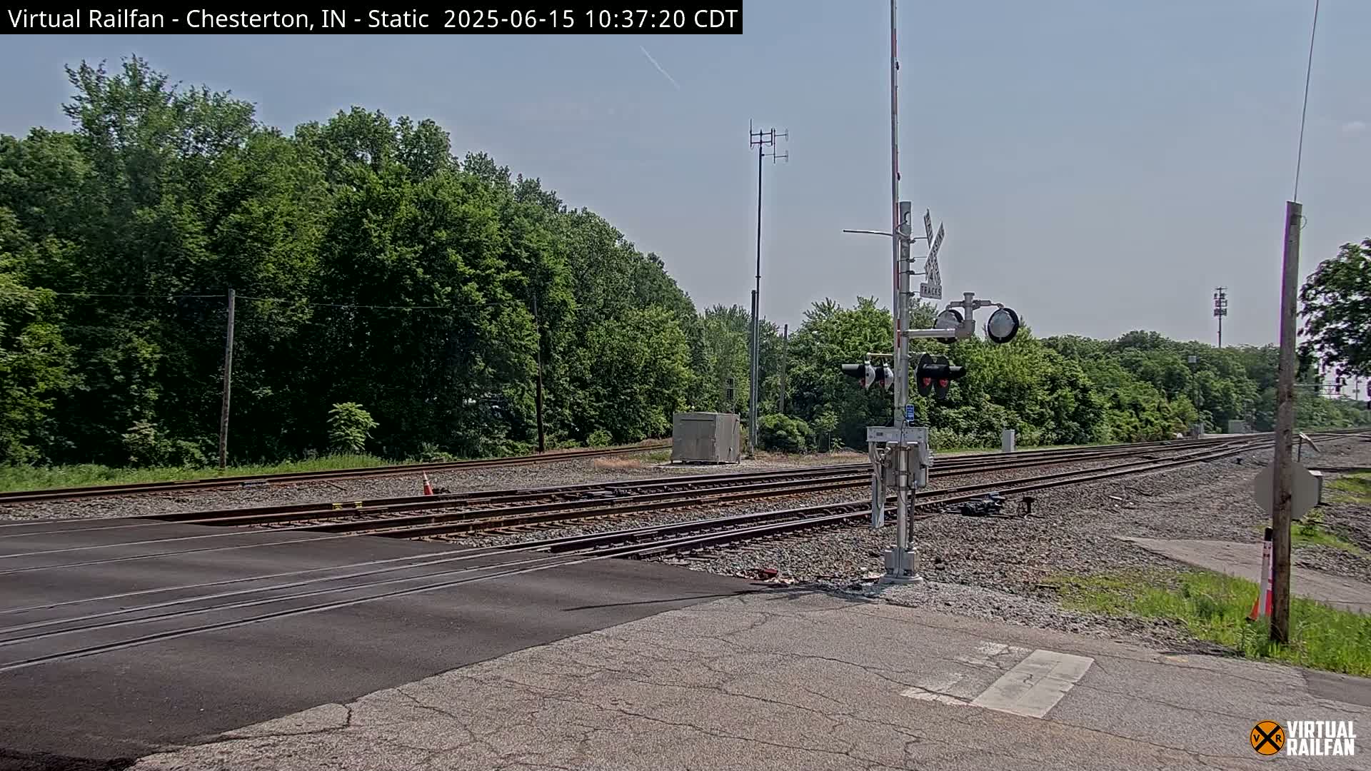 A railroad crossing with multiple tracks intersects a road, surrounded by lush green trees under a clear, sunny sky.