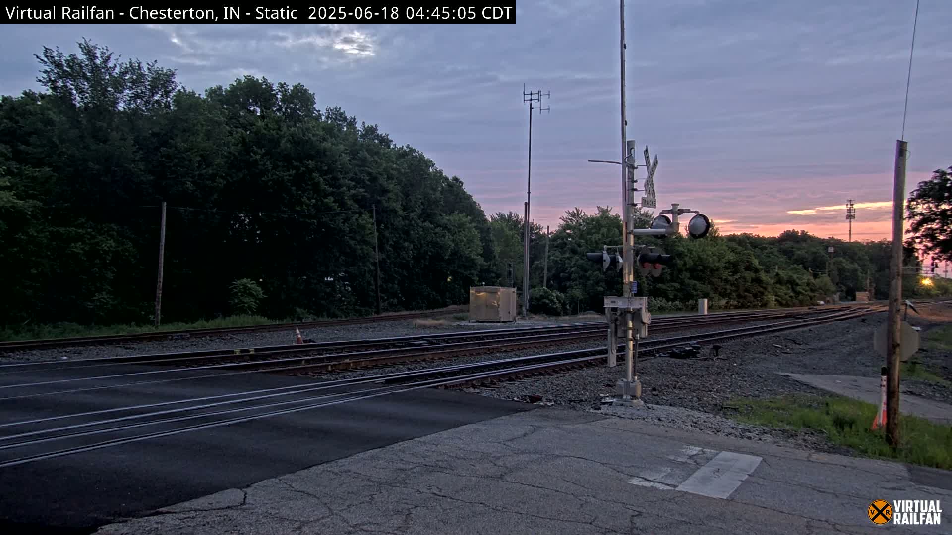A railroad crossing at dawn under a mostly clear, pinkish sky, with several sets of train tracks intersecting a road and surrounded by lush green trees.