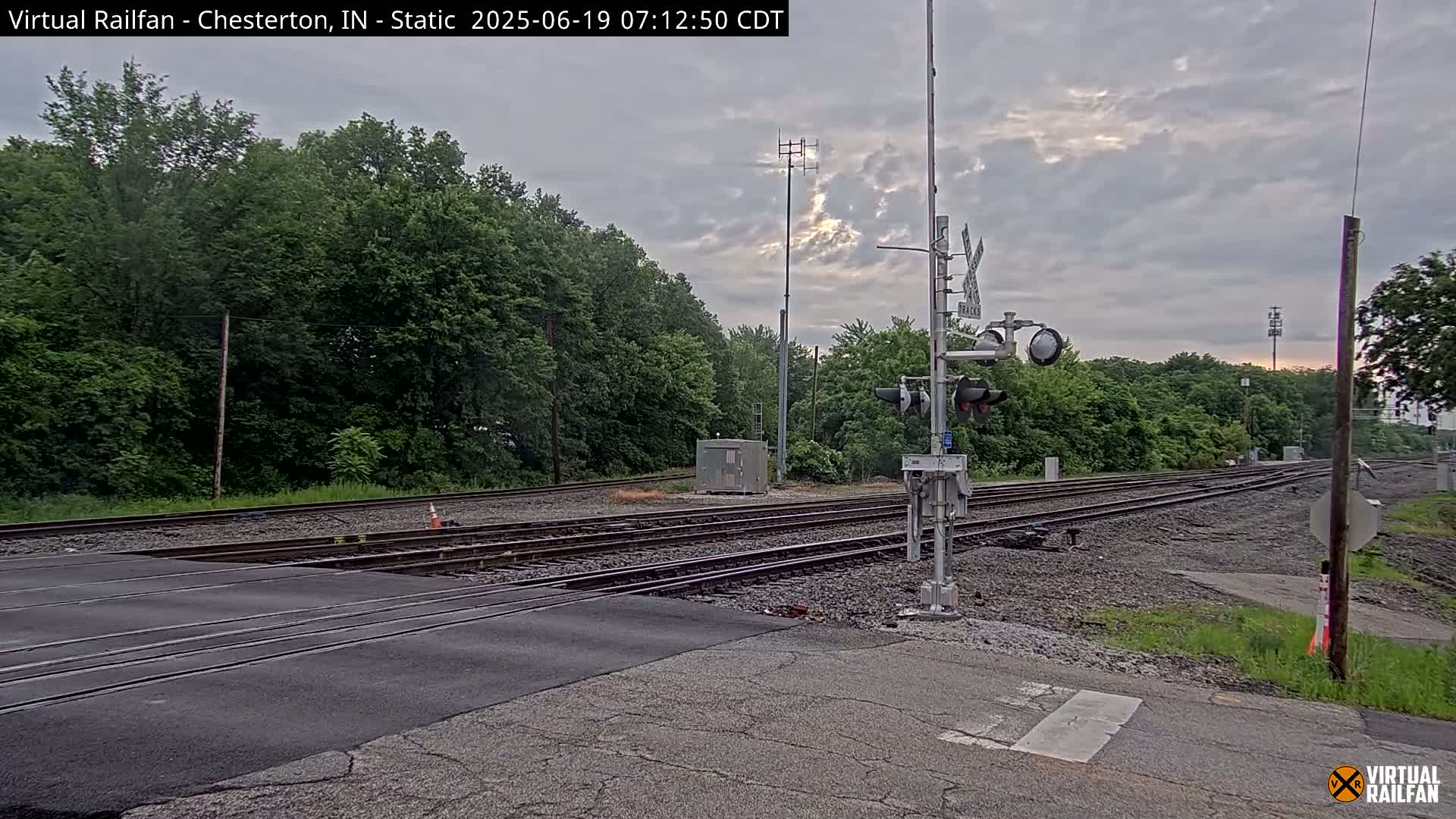 A railroad crossing with train tracks intersecting a paved road, surrounded by lush green trees under an overcast sky.