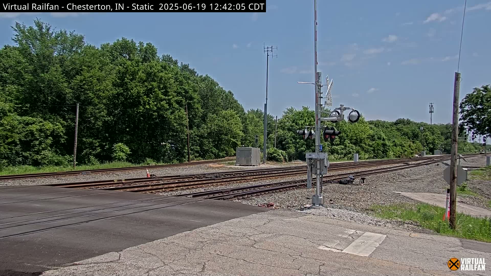 A railroad crossing with train tracks and a signal system is shown on a sunny day, surrounded by lush green trees.