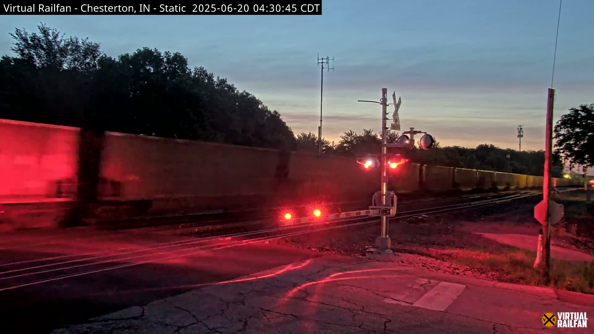 A long freight train passes a railroad crossing at twilight under a clear sky, illuminated by the red lights of the crossing gates.
