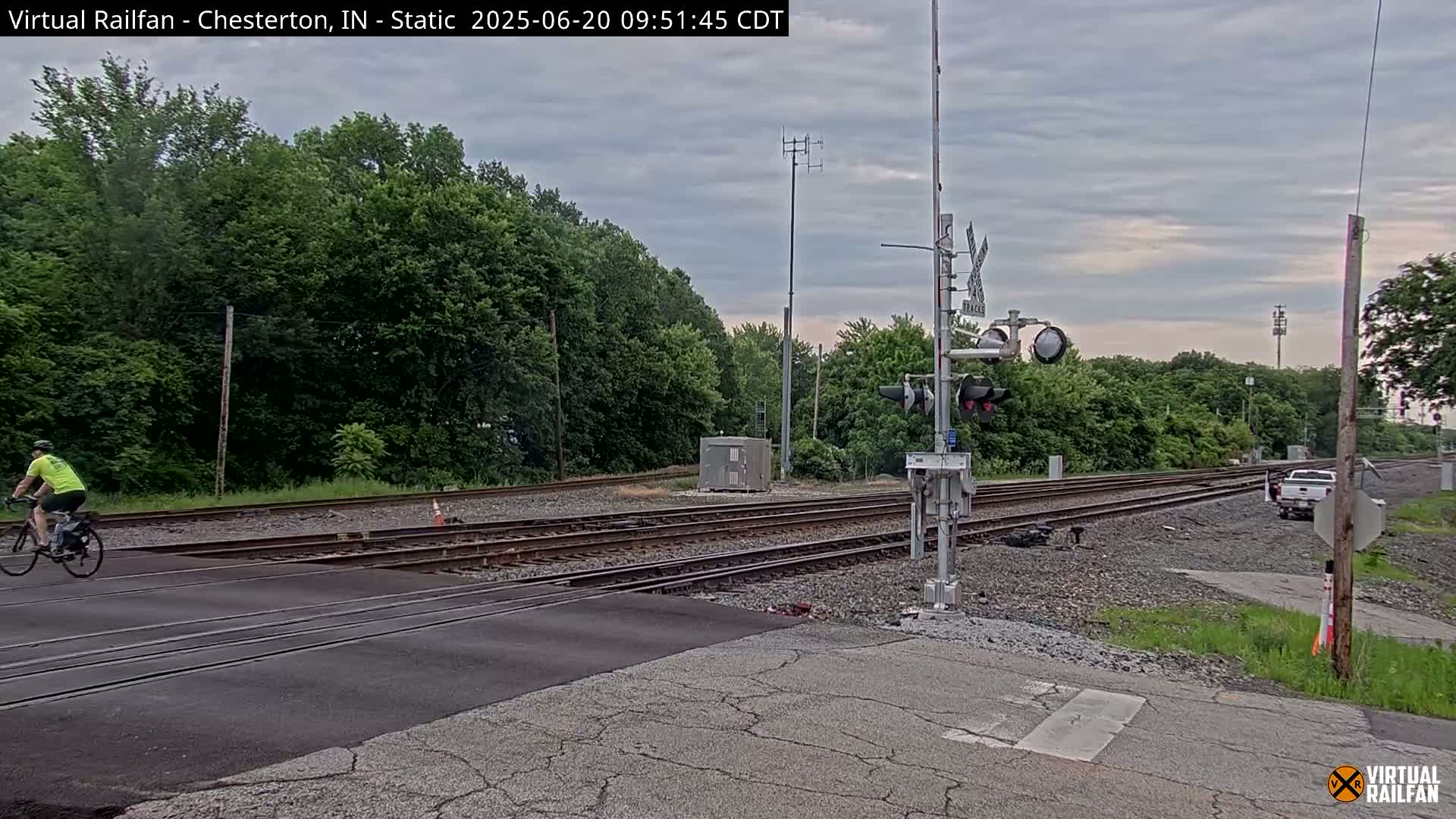 A cyclist in a bright yellow shirt rides a bicycle across a road and railroad crossing under an overcast sky, past a white truck and railroad signals.