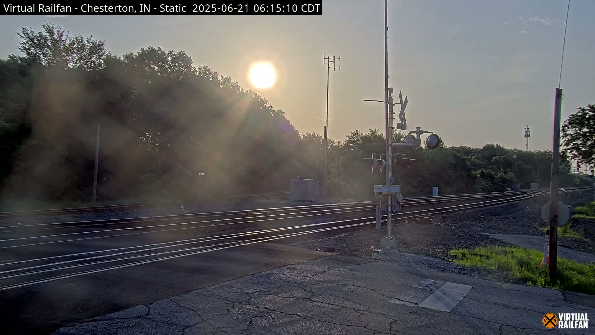 A railroad crossing is shown at sunrise, with multiple train tracks intersecting a road, and trees and other vegetation surrounding the scene.