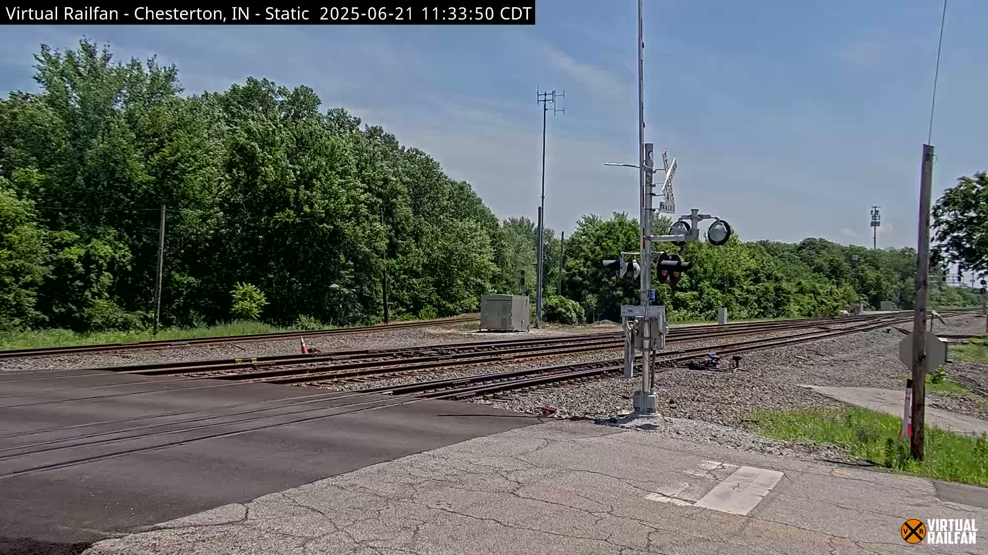 A railroad crossing with multiple tracks is seen on a sunny day, bordered by lush green trees.