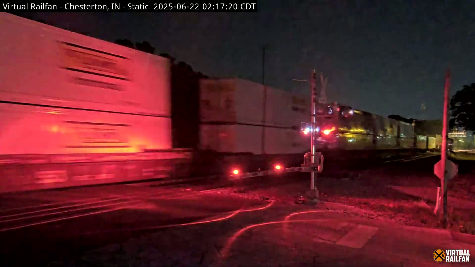 A long freight train passes through a railroad crossing at night, illuminated by the red lights of the crossing gates.