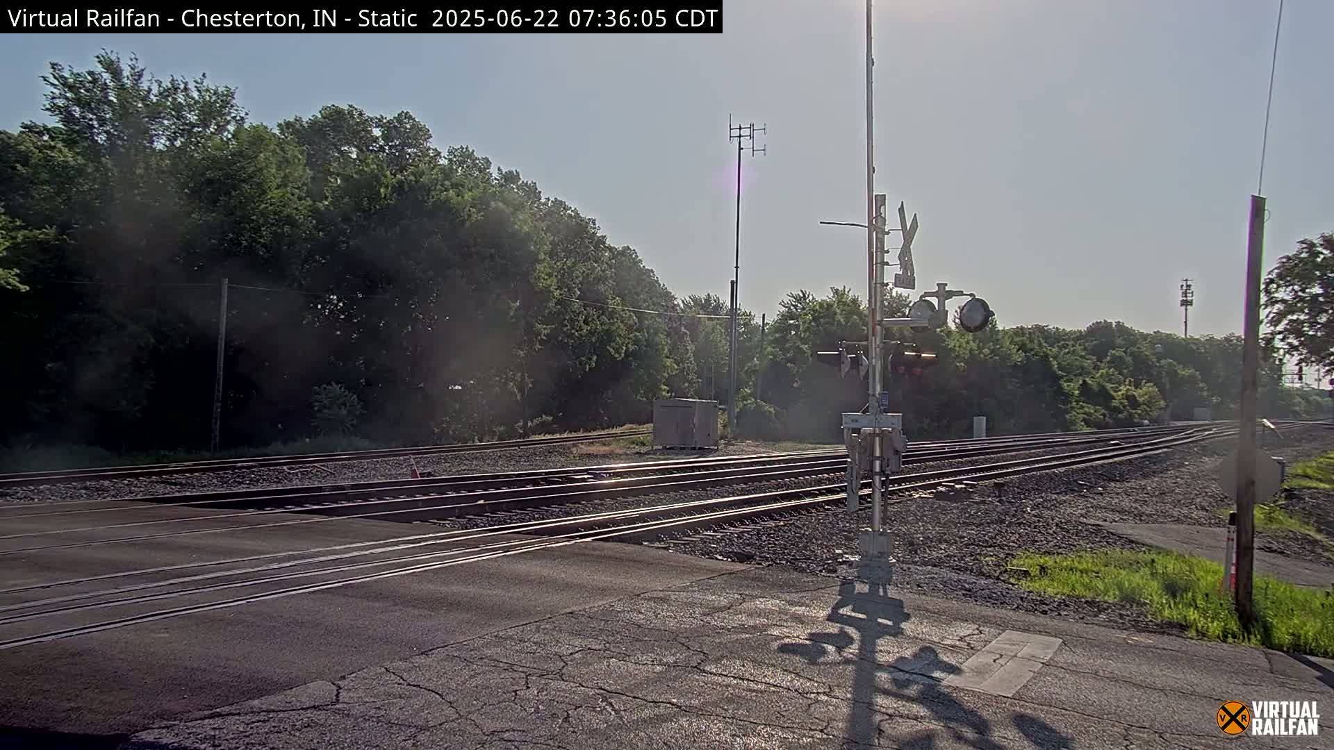 A sunny day at a railroad crossing shows train tracks intersecting a road, with green trees and vegetation in the background.