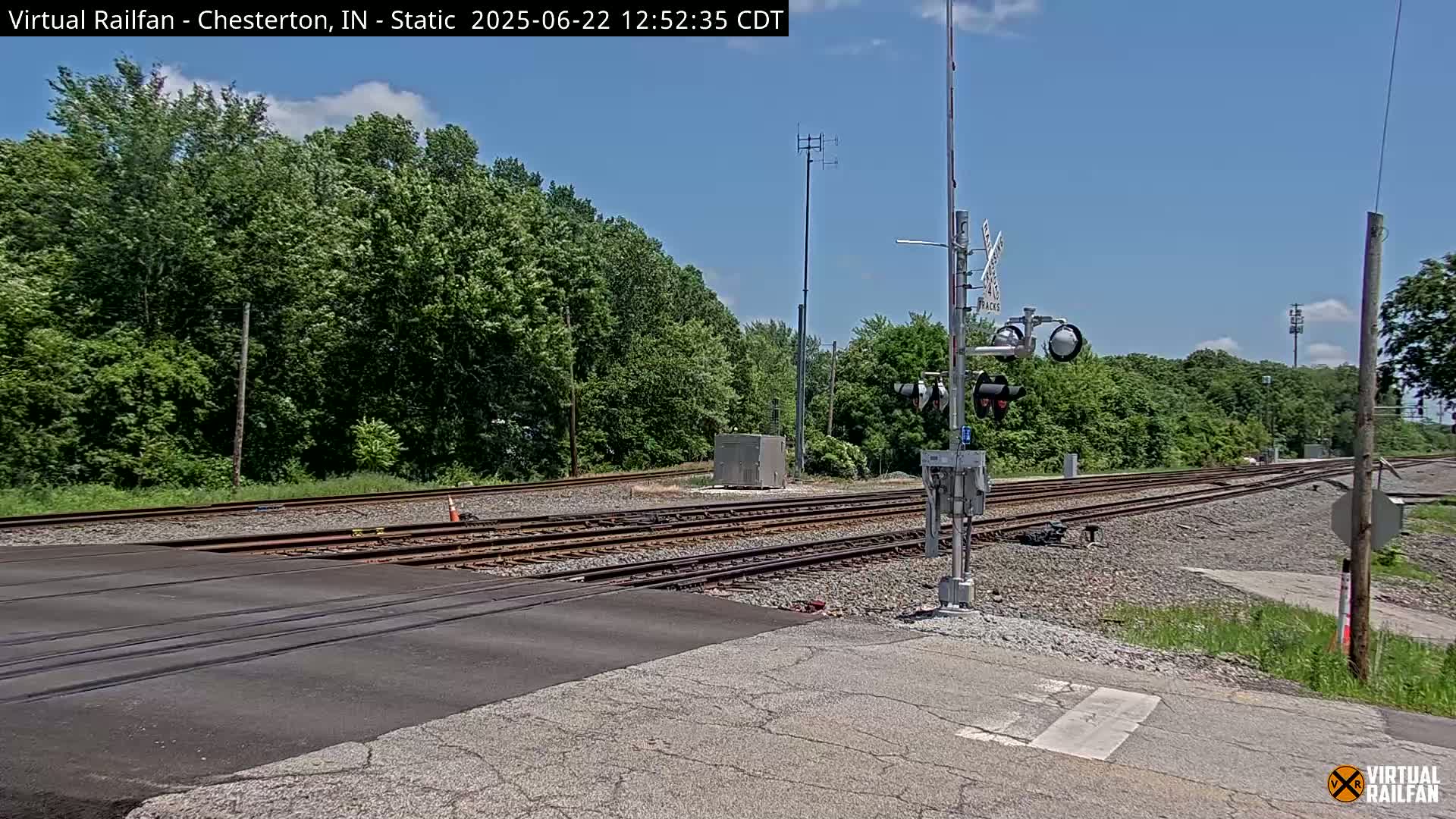 A railroad crossing with multiple train tracks is shown on a sunny day, bordered by lush green trees.