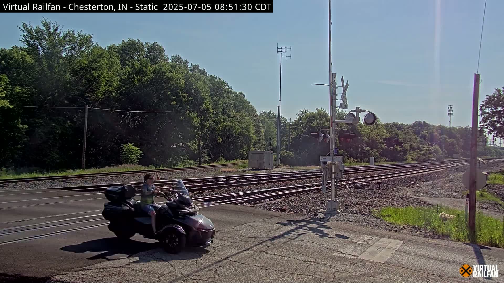 A person sits on a three-wheeled motorcycle at a railroad crossing on a sunny day.