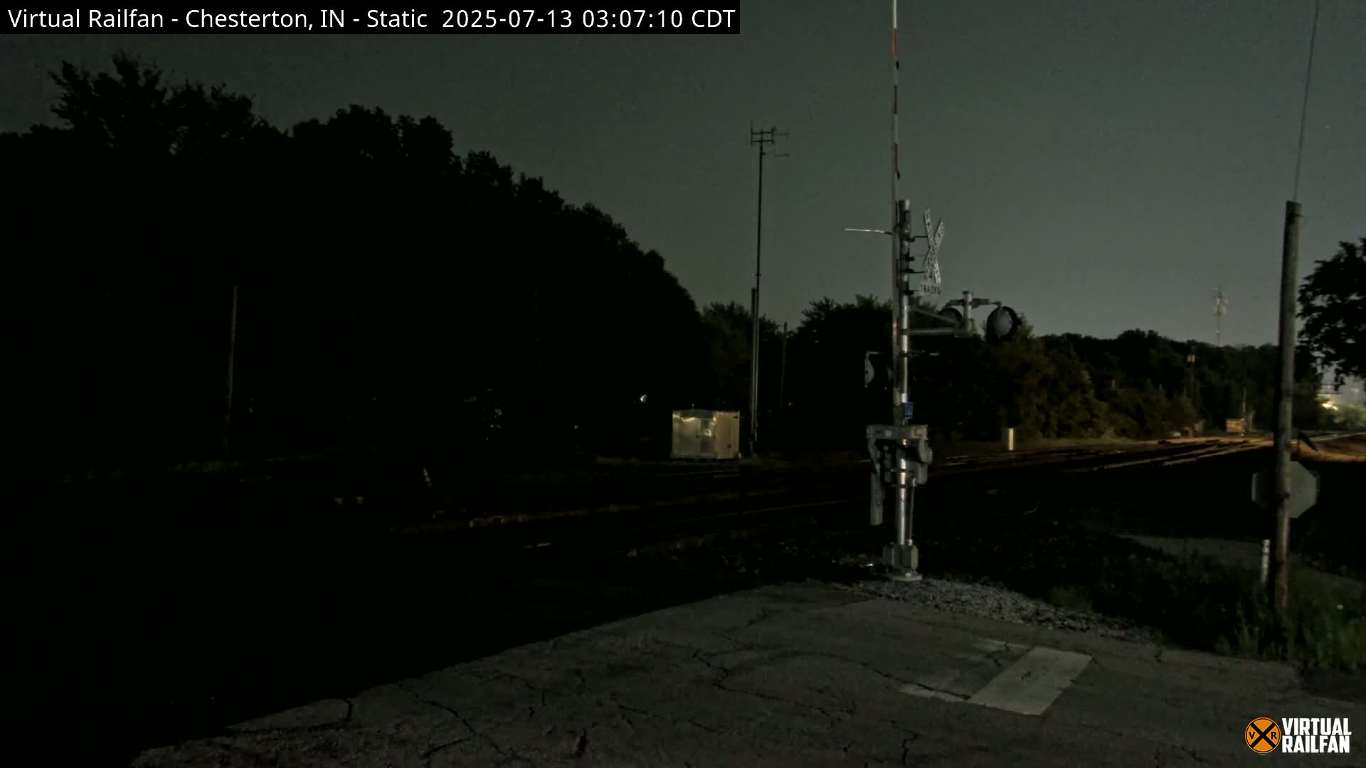 A nighttime view of railroad tracks and crossing signals, surrounded by dark trees under a cloudy sky.