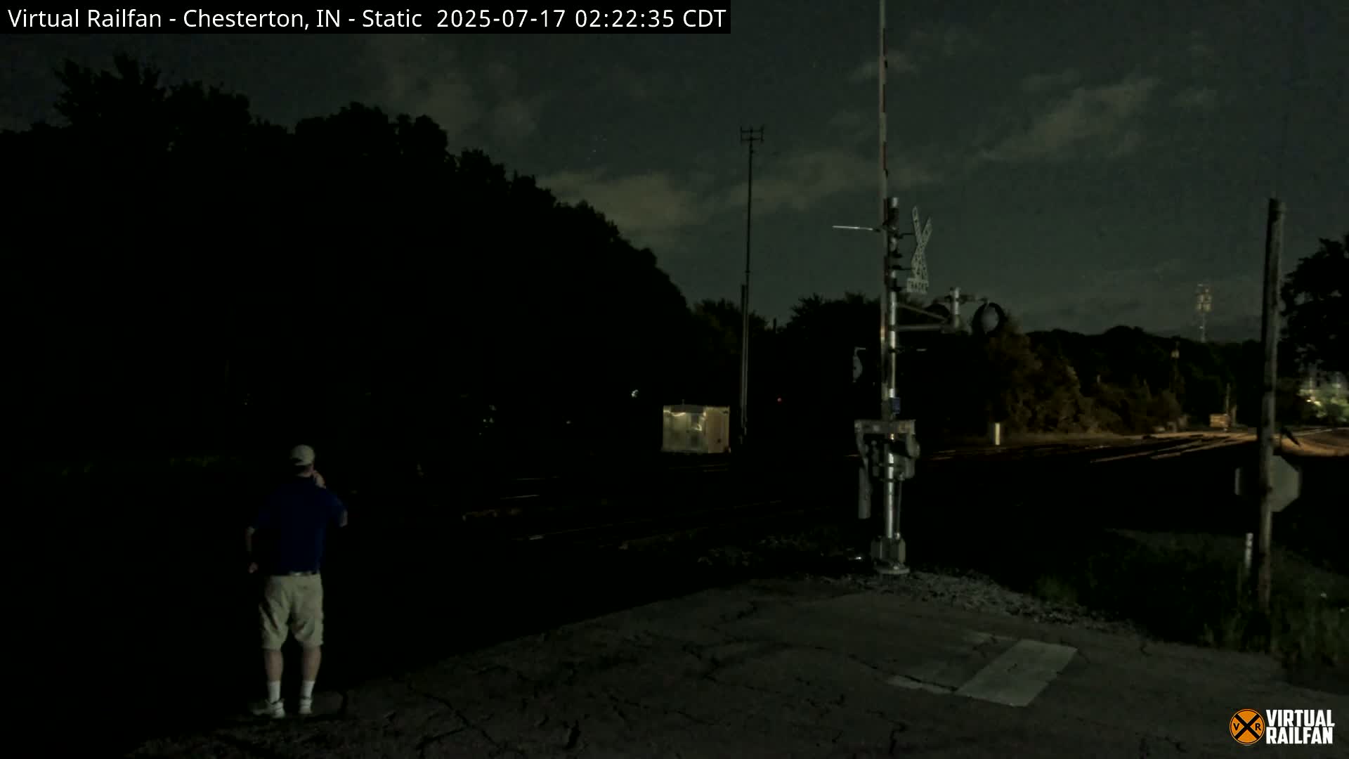 A man stands near railroad tracks at night under a partly cloudy sky.