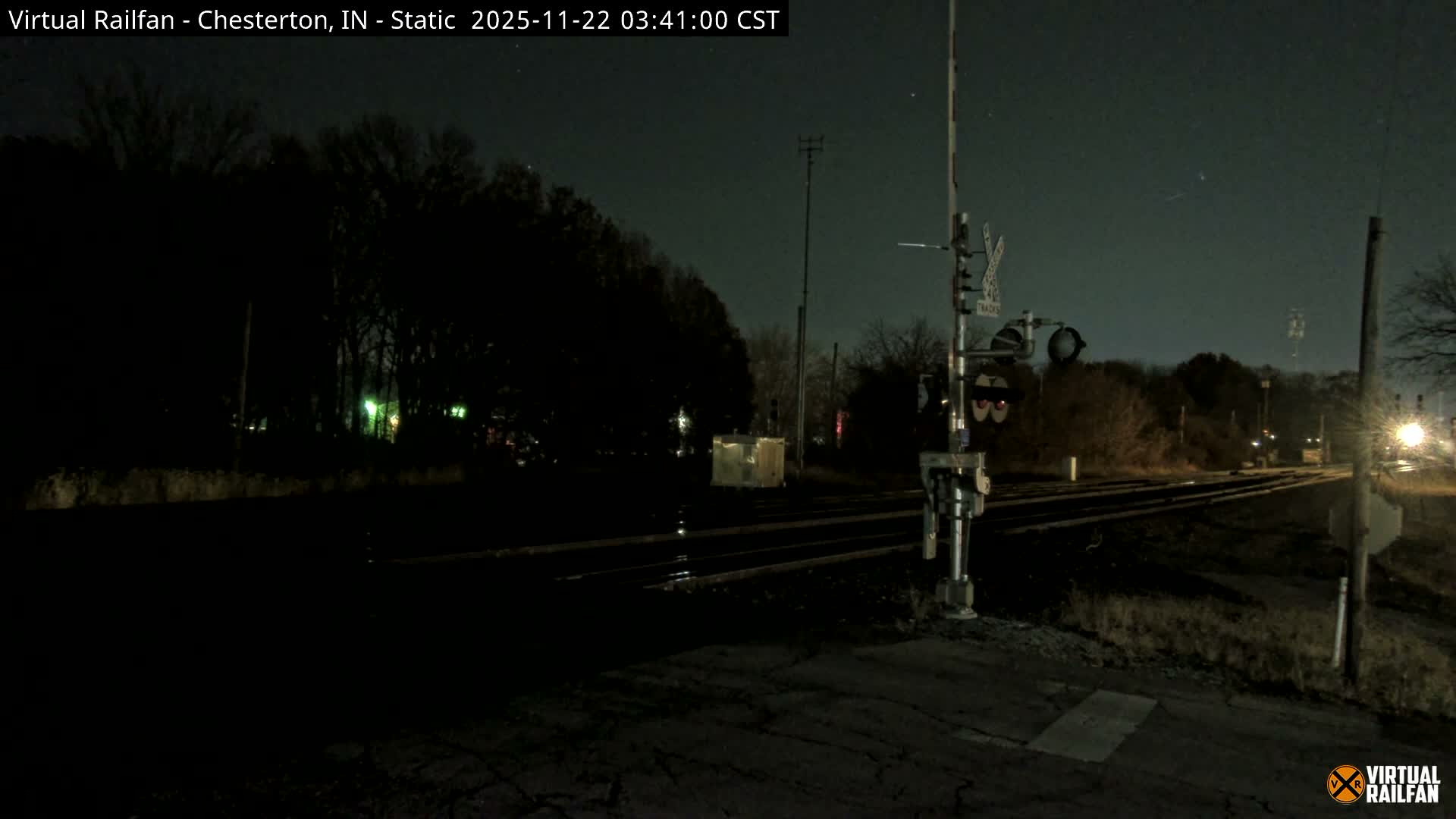 The image shows a dark outdoor scene at a railway crossing with multiple tracks, a signal post with a crossbuck sign, and distant lights under clear, dry night conditions.