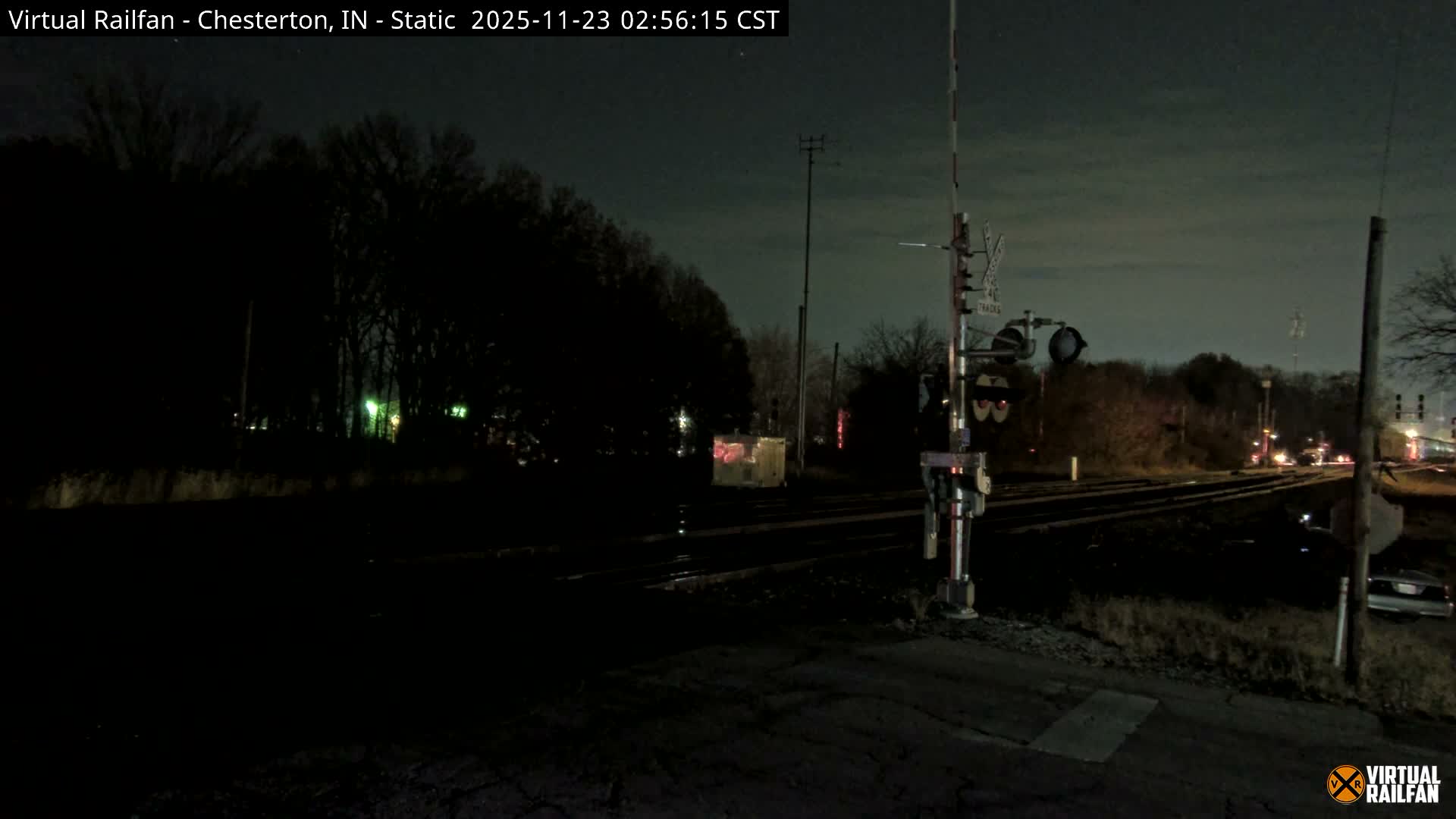 A railroad crossing with multiple tracks, a signal gate, and a nearby shed is visible at night under a clear or partly cloudy sky, bordered by dark trees and distant artificial lights.