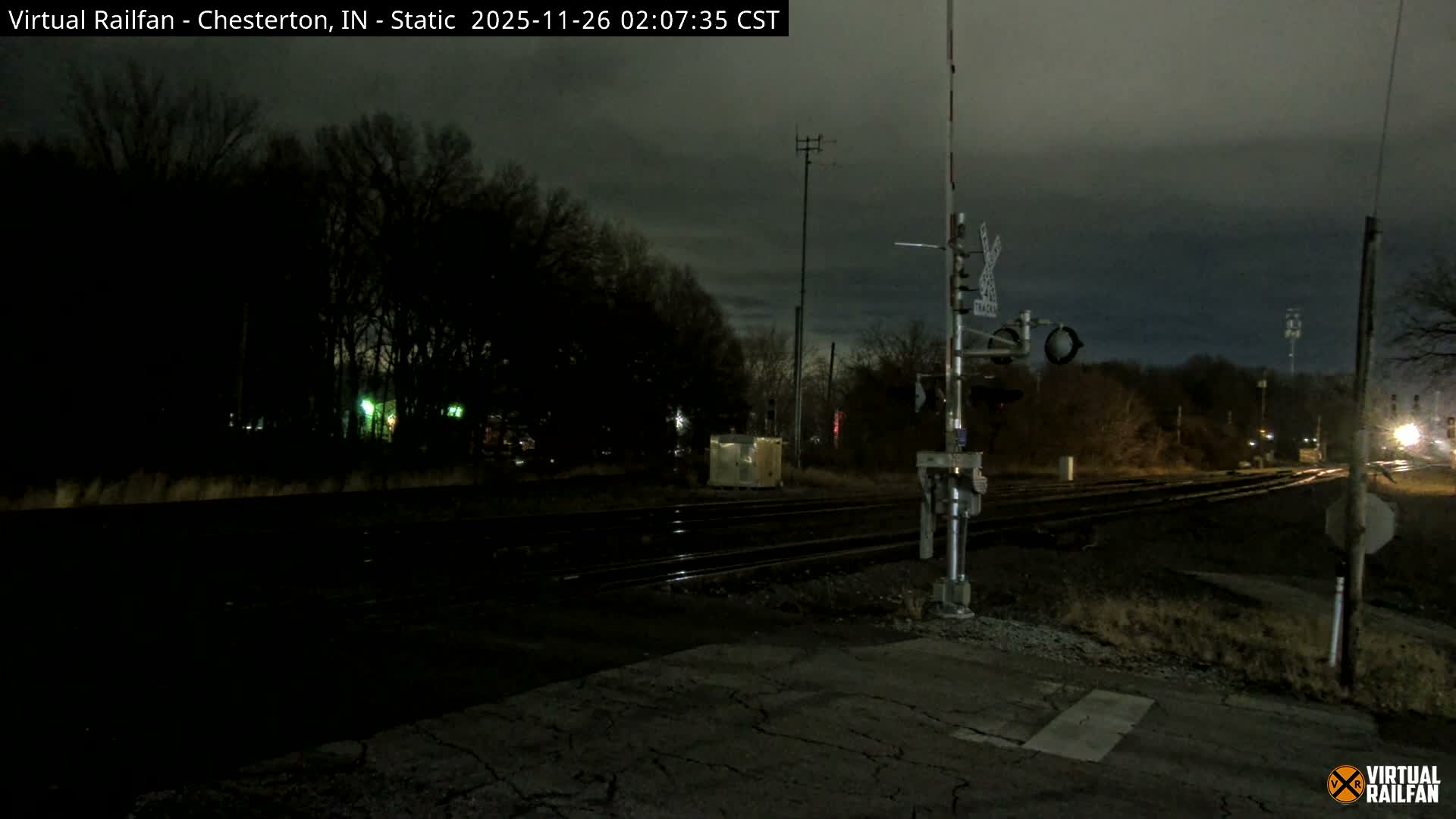 The image captures a dark, overcast night at a multi-track railroad crossing with a raised gate and signal, bordered by dense trees and distant lights.
