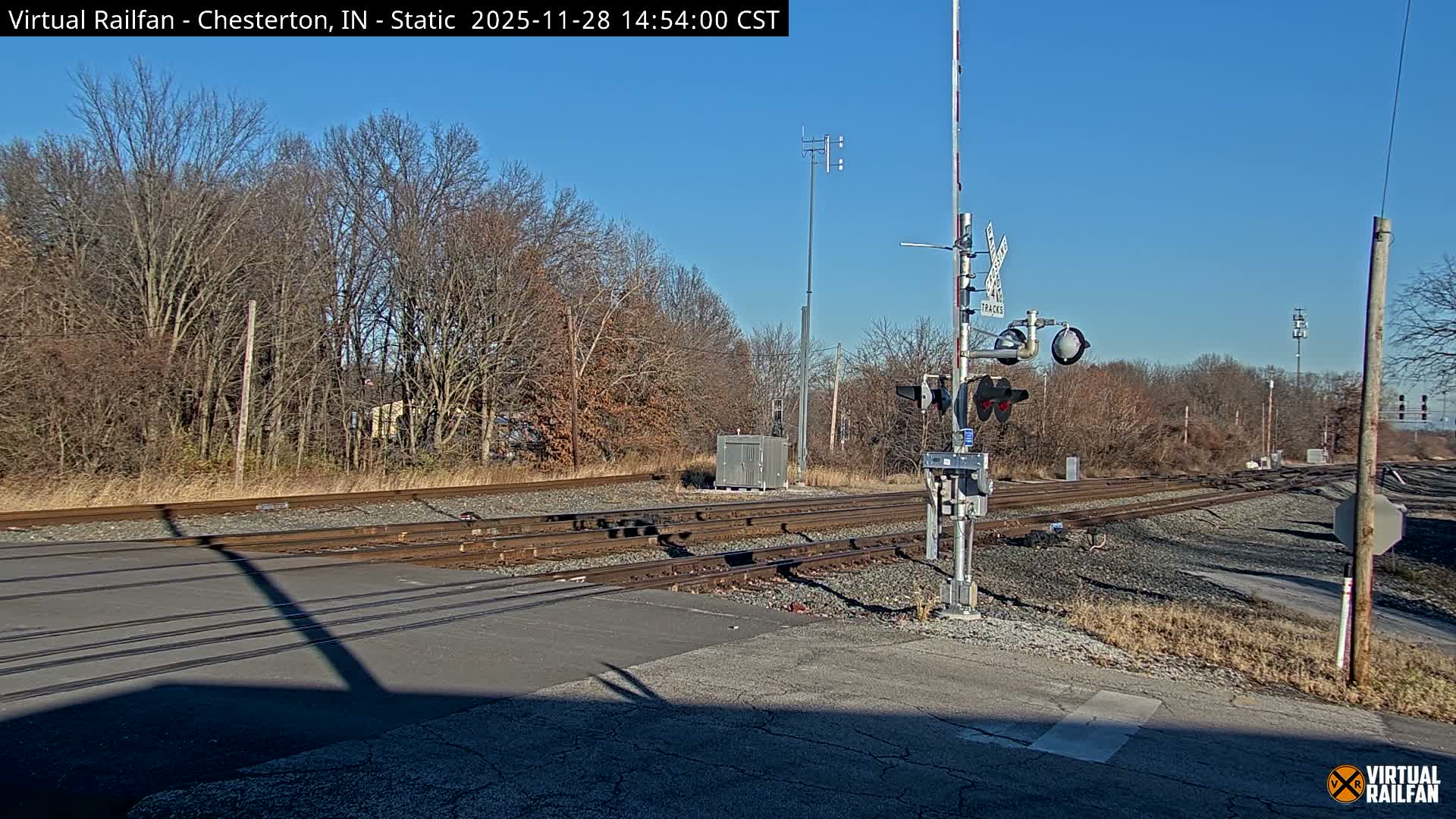 A railroad crossing with multiple tracks over a paved road is depicted on a clear, sunny day, featuring a raised crossing gate and signal in the foreground, and dormant trees lining the background.