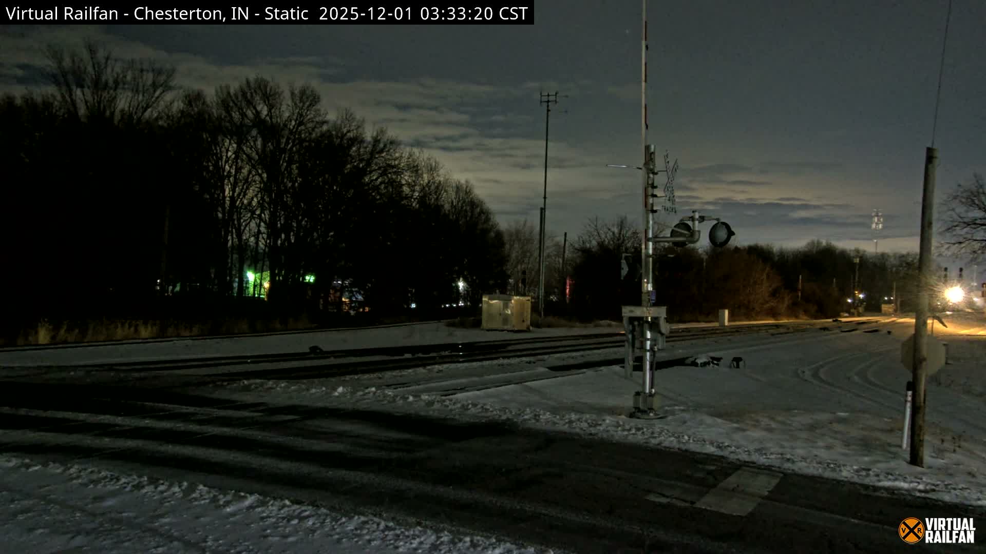 The image shows a snowy railroad crossing at night under a cloudy sky, with dark trees in the background and distant artificial lights.