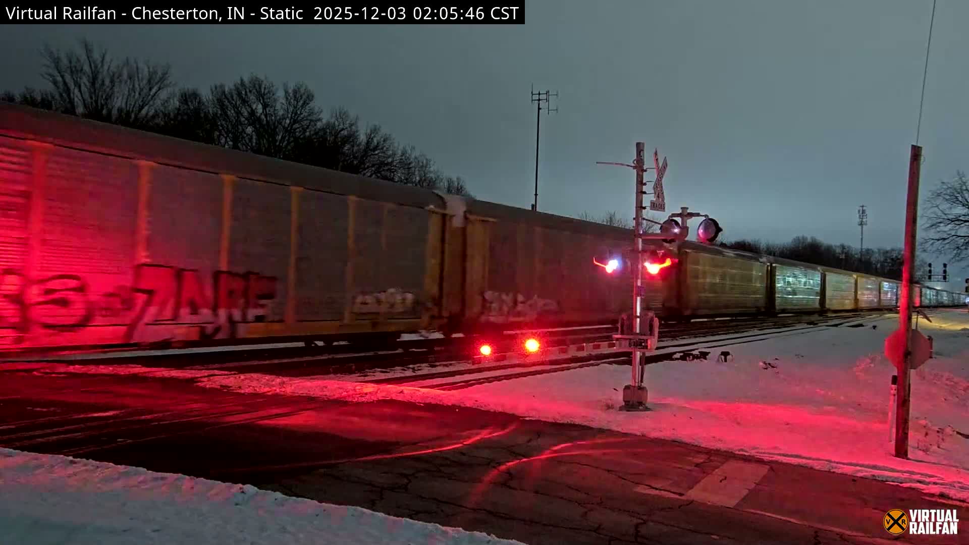 A freight train, illuminated by the flashing red lights of a railroad crossing, moves through a snow-covered landscape under a dark, wintry sky.