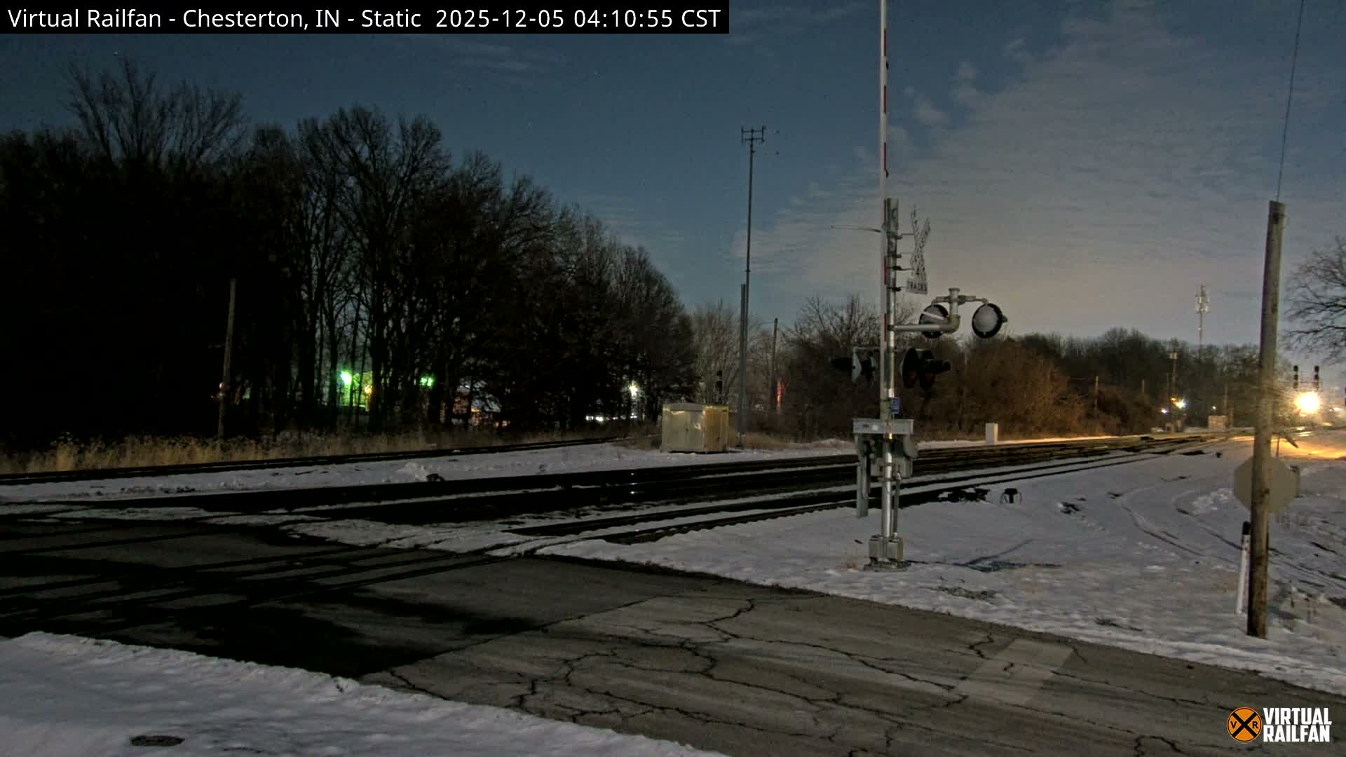 A snow-covered railway crossing with multiple tracks and a lowered barrier is seen under a partially cloudy night sky, with a distant bright light on the tracks to the right.