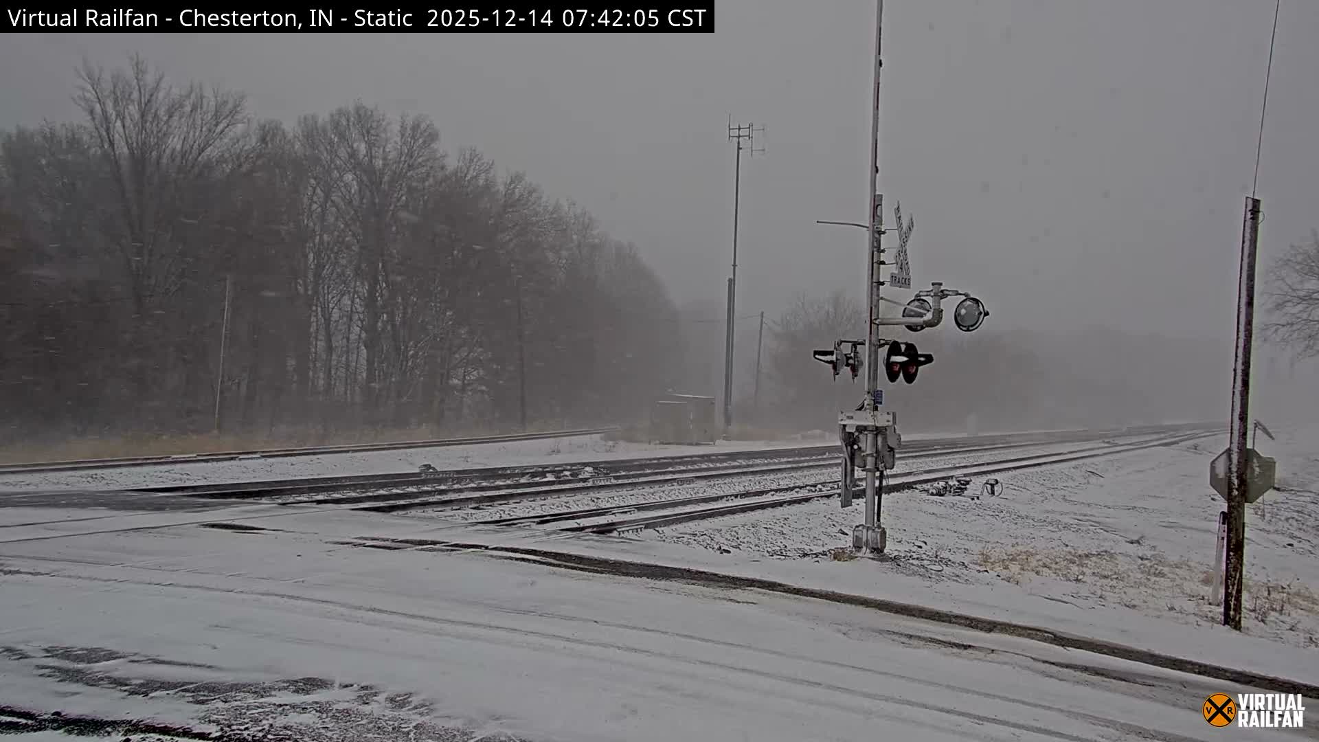 A snow-covered railway crossing with multiple tracks and a lowered barrier is seen under a partially cloudy night sky, with a distant bright light on the tracks to the right.