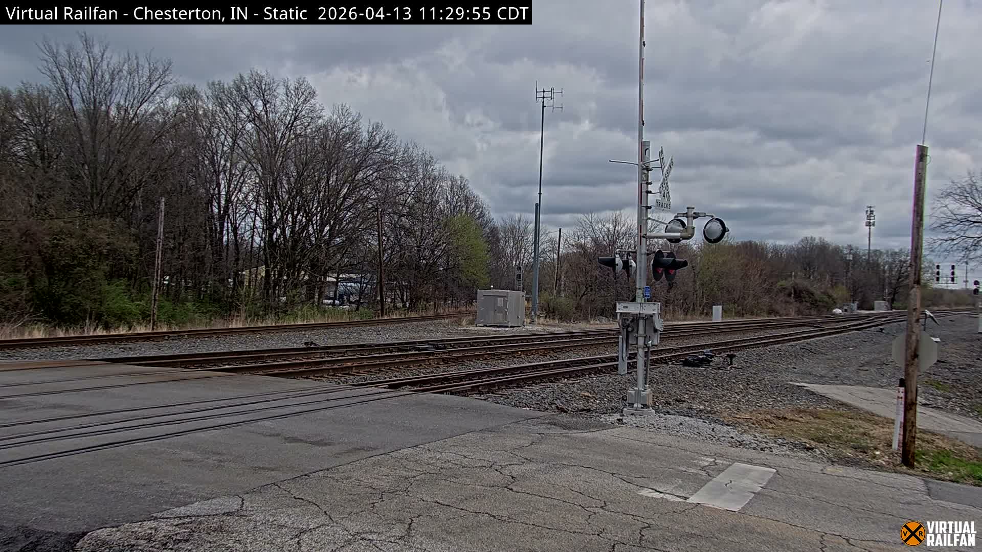 A snow-covered railway crossing with multiple tracks and a lowered barrier is seen under a partially cloudy night sky, with a distant bright light on the tracks to the right.