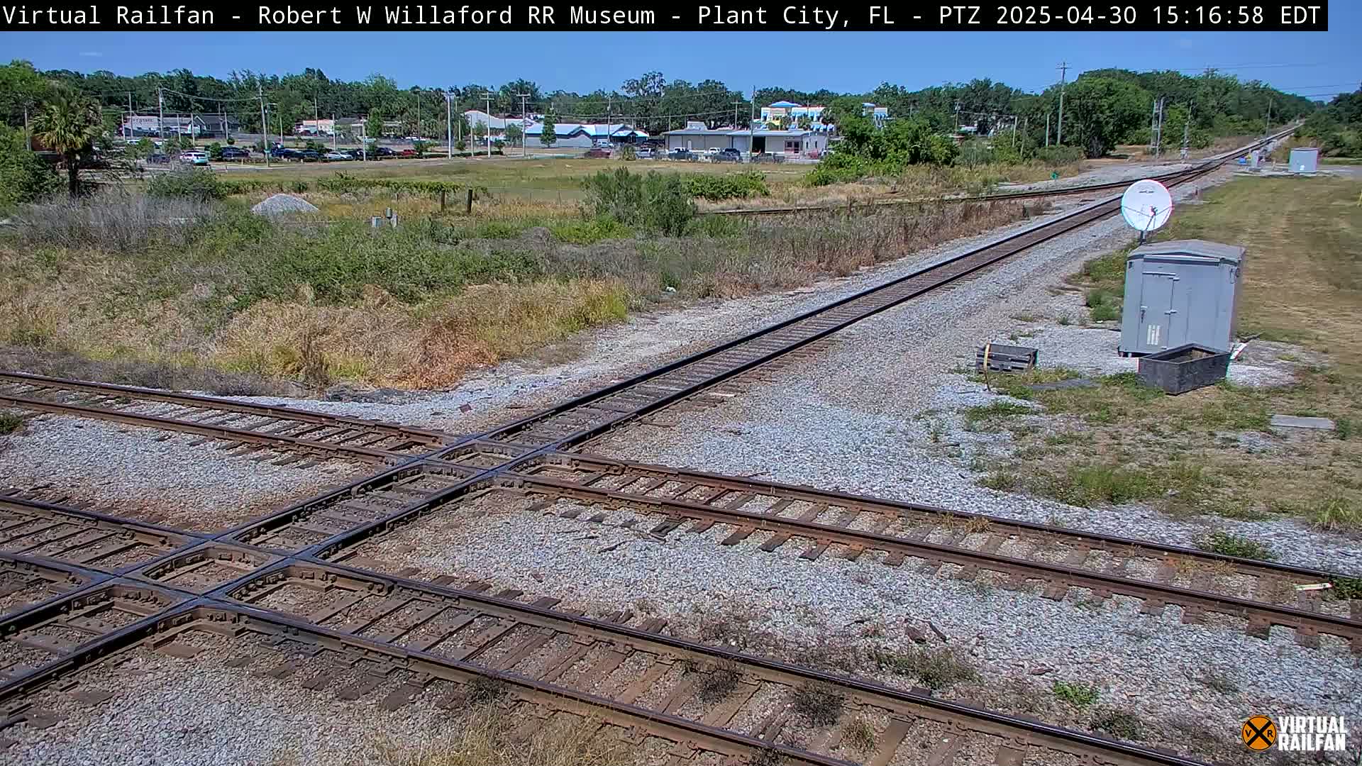 A sunny day shows a railroad crossing with two sets of tracks intersecting, surrounded by sparse vegetation and a small building near a satellite dish.