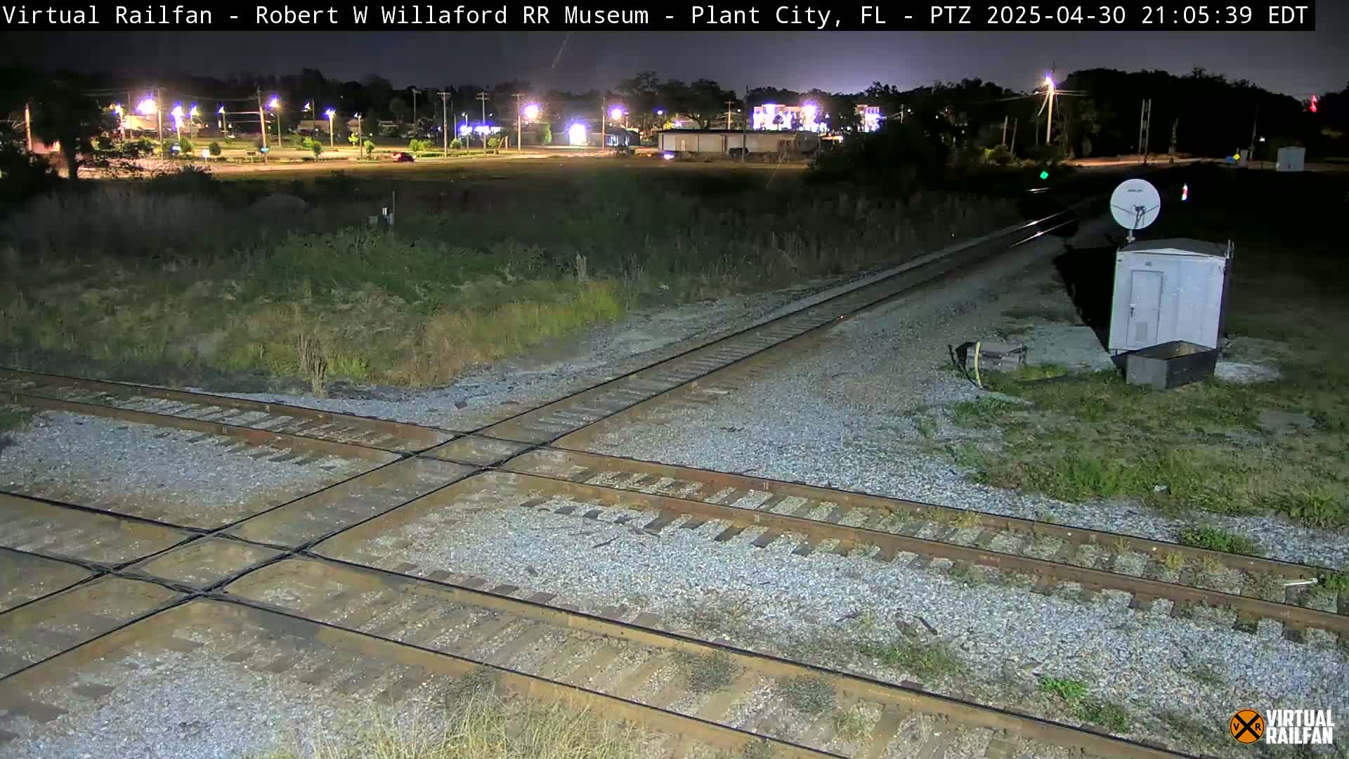 A nighttime view of a railroad crossing with intersecting tracks, a small building, and a satellite dish, surrounded by sparse vegetation under a clear sky.