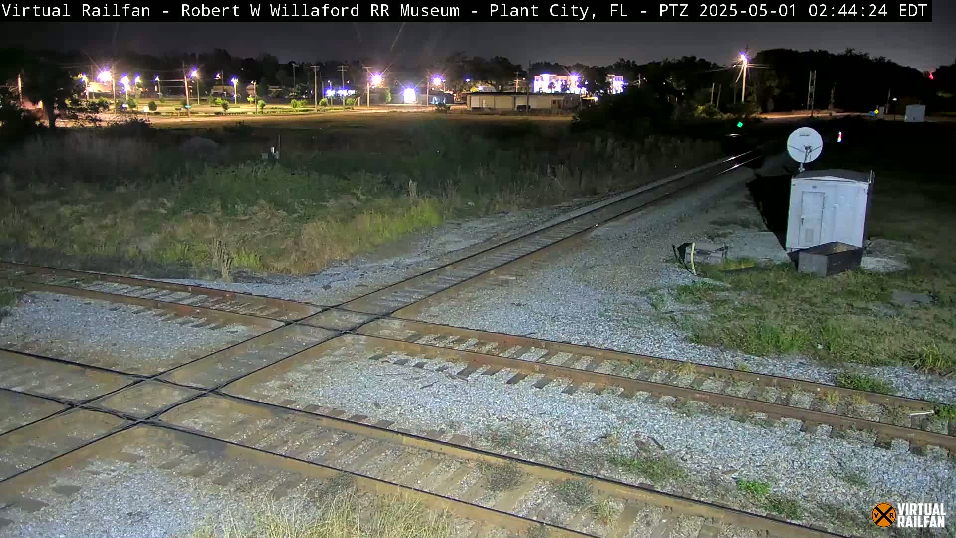 A nighttime view of a railroad crossing with a small building and satellite dish nearby, under clear skies.