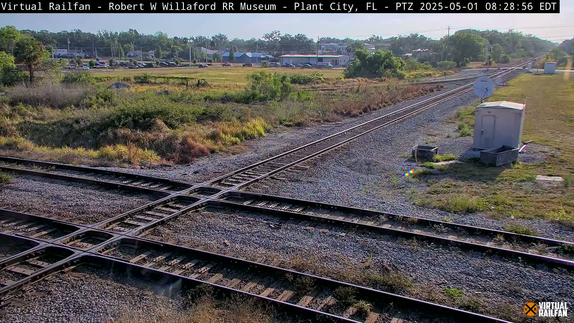 A sunny outdoor view of a railroad crossing with two sets of tracks intersecting, surrounded by scrubby vegetation and a small utility building.