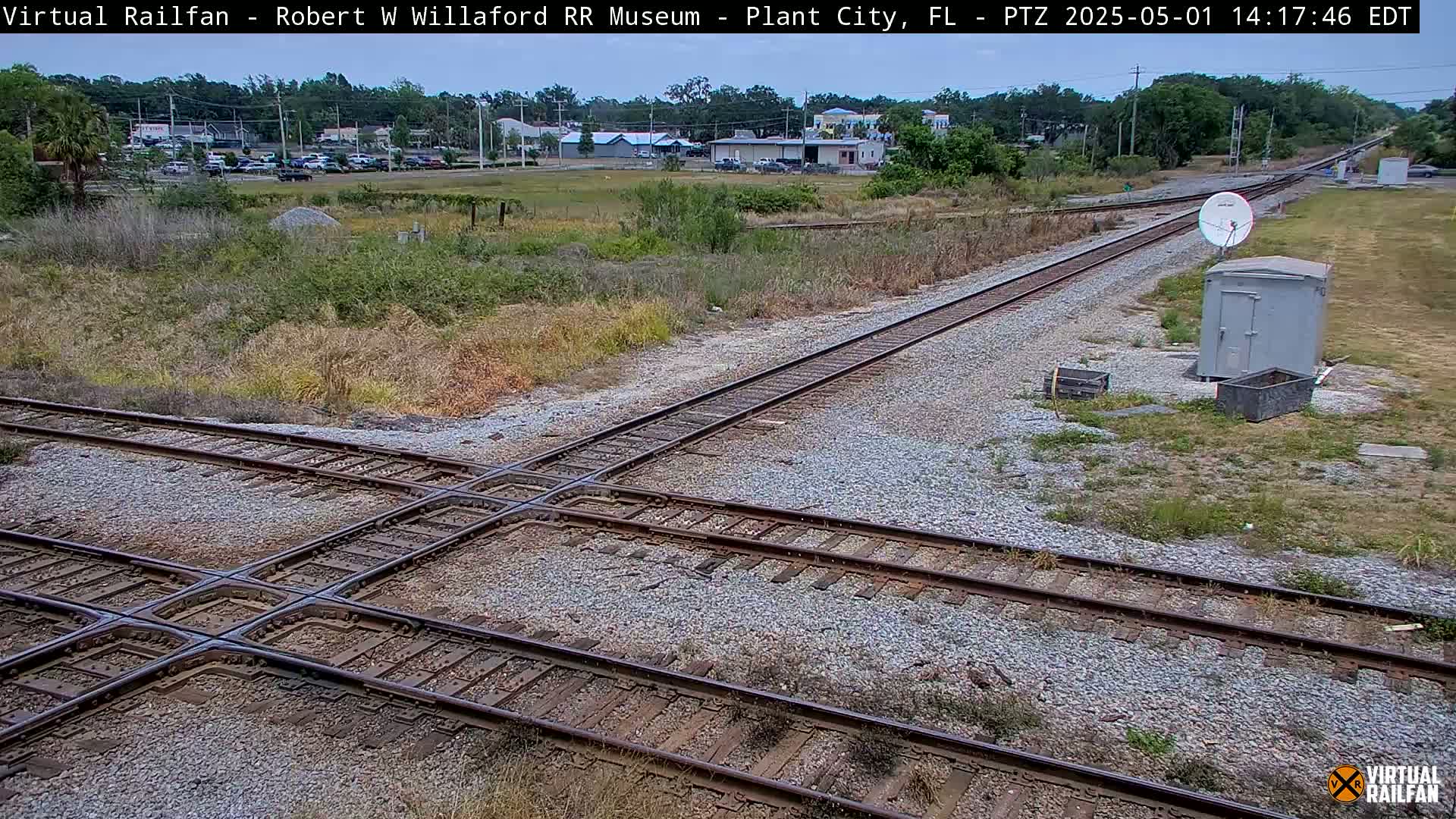 A railroad crossing with two sets of tracks intersects, surrounded by scrubby vegetation under a partly cloudy sky.