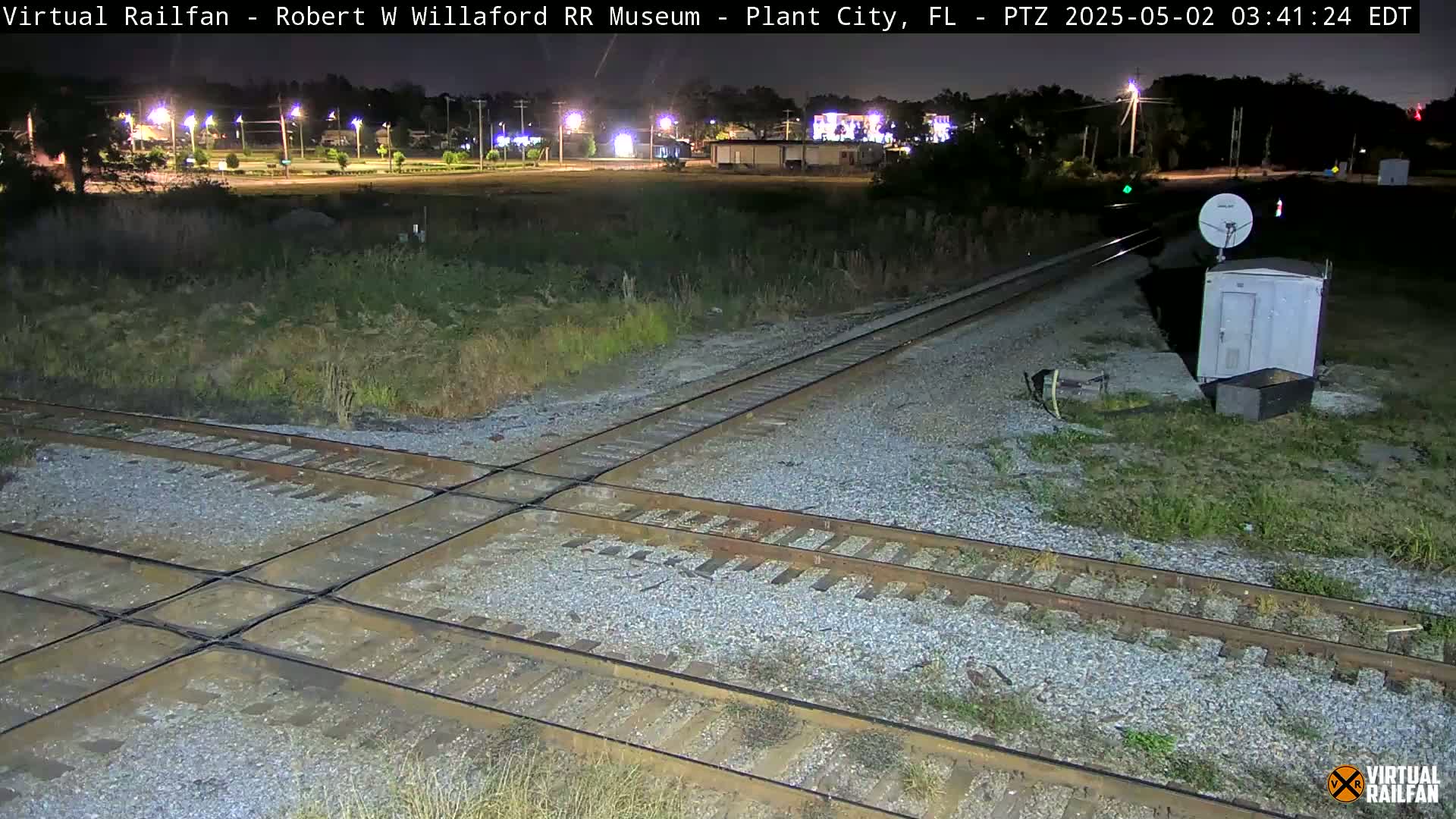 A nighttime view of a railroad crossing with two sets of tracks intersecting, a small white utility box nearby, and a sparsely vegetated area surrounding the tracks.