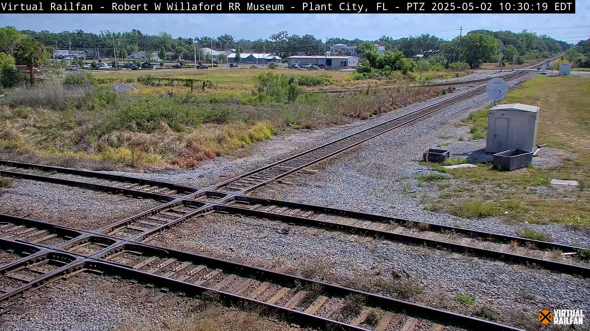 A sunny outdoor scene shows a railroad crossing with two sets of tracks intersecting, surrounded by scrubby vegetation and a small utility building.