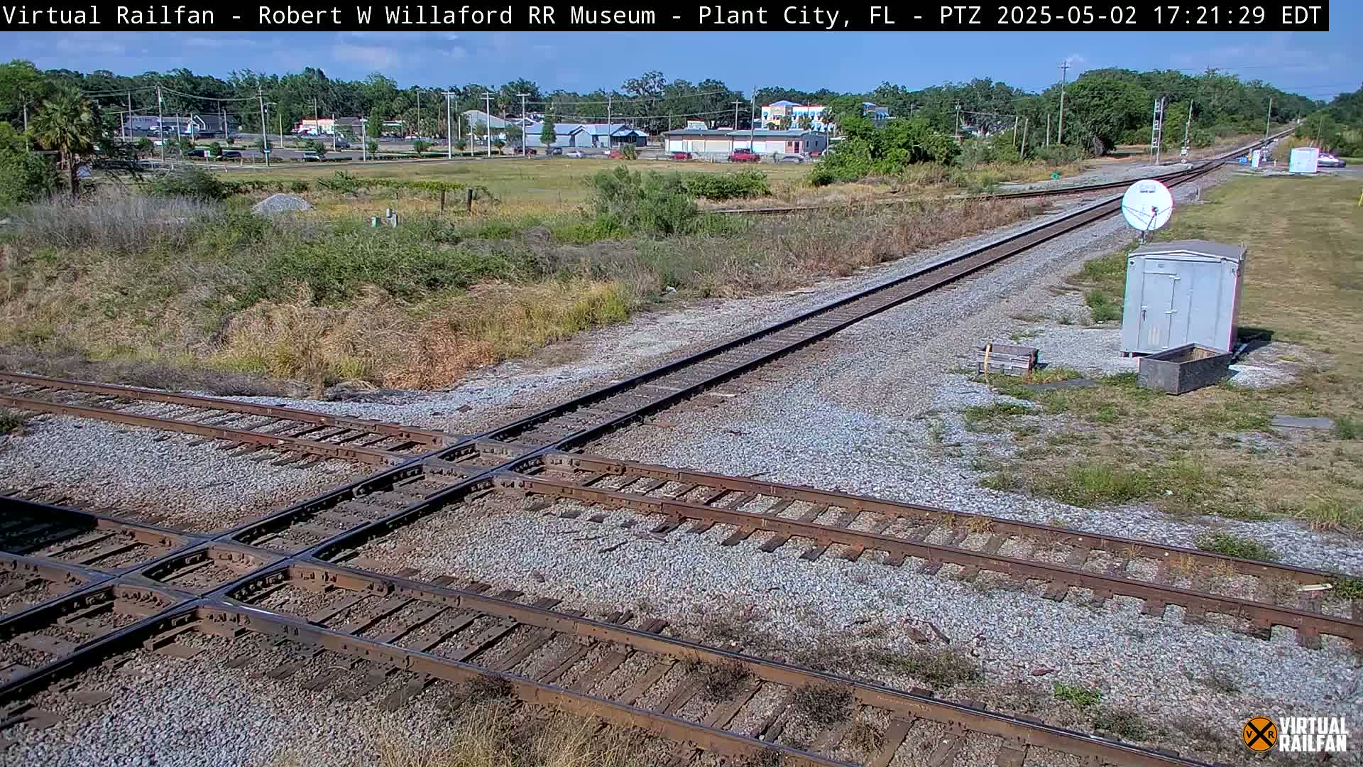 A sunny outdoor scene shows a railroad crossing with two sets of tracks intersecting, a small gray shed and satellite dish nearby, and buildings in the distance.