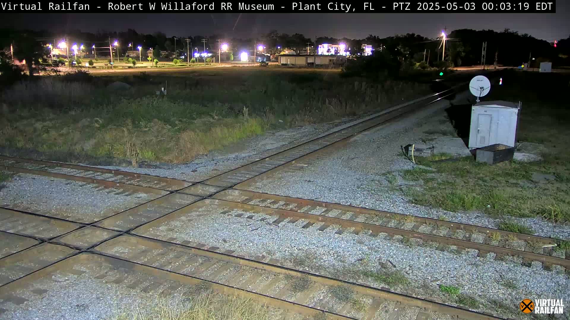 A nighttime view of a railroad crossing with a small building and satellite dish nearby, under clear skies.