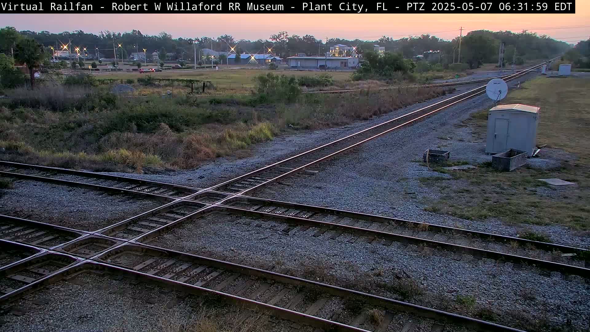 Two sets of train tracks intersect at a railroad crossing near a small building under a twilight sky.