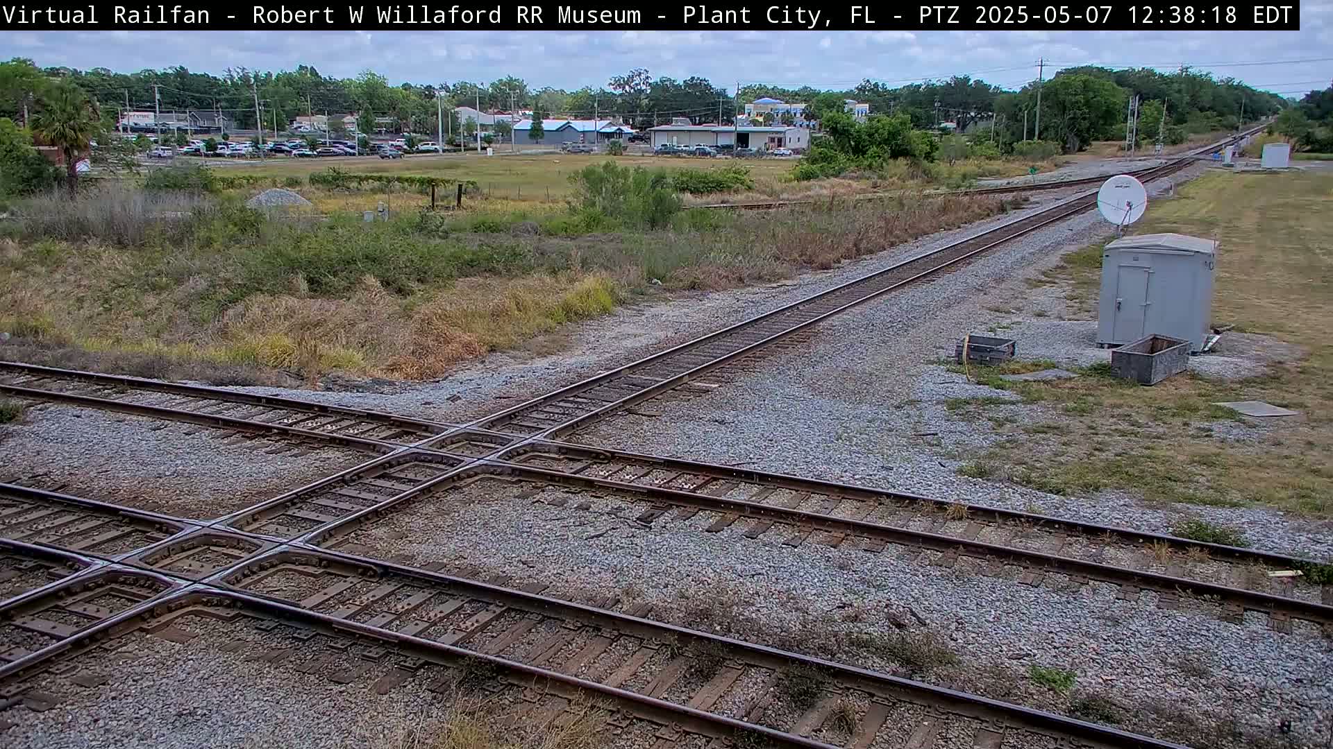 A high-angle view shows a railroad crossing with two sets of tracks intersecting, surrounded by dry, grassy terrain and a small, gray utility shed under a mostly clear sky.