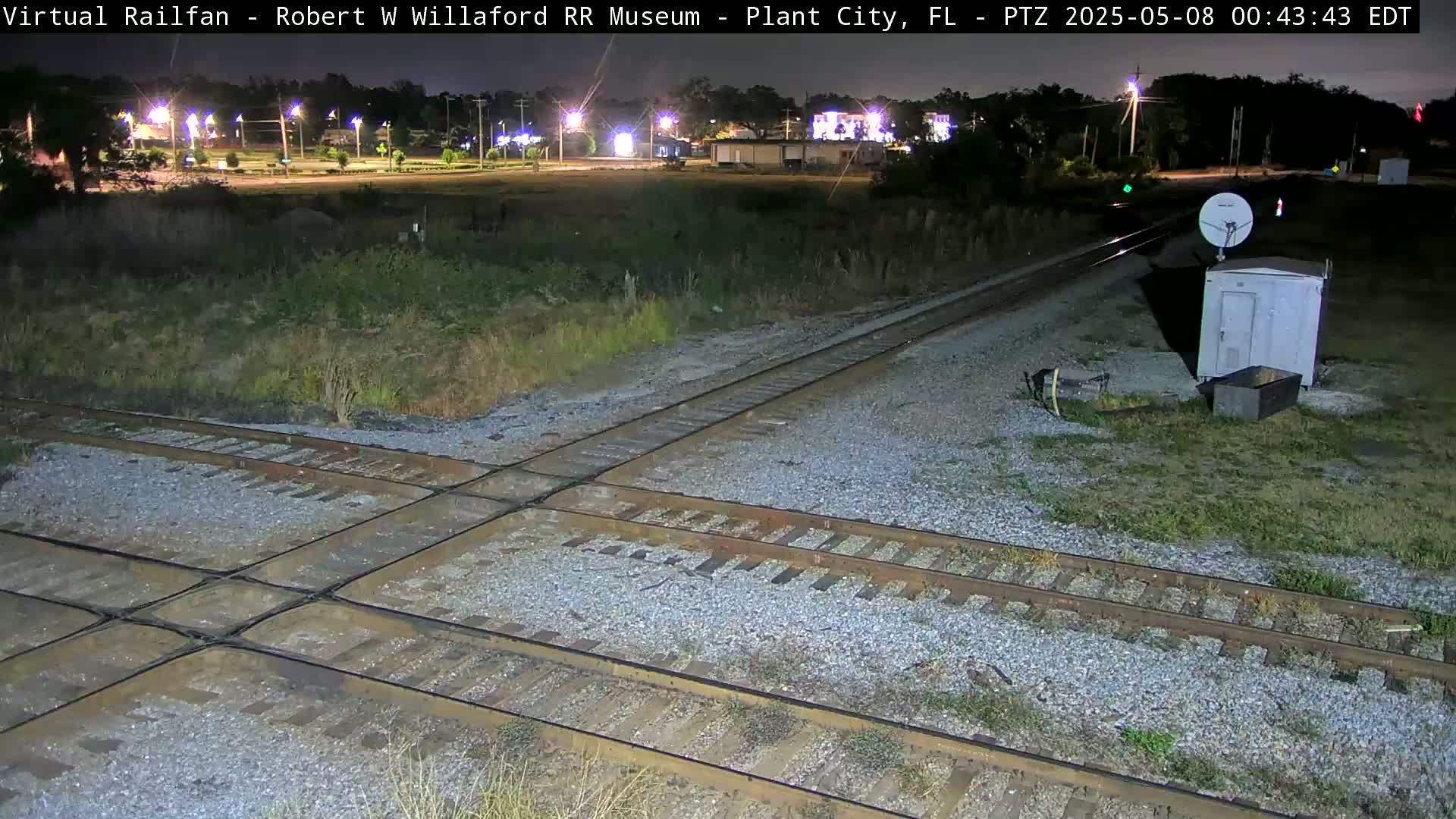 A nighttime view of a railroad crossing with a small building and satellite dish, under a clear sky.