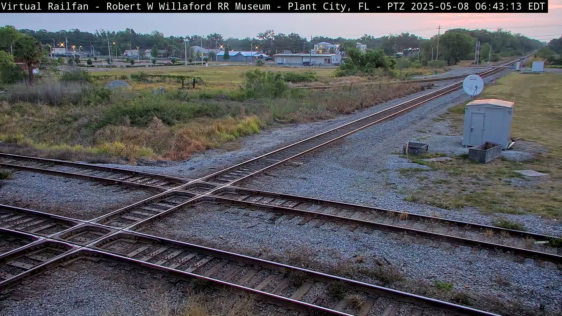 A high-angle, long shot shows a railroad crossing under a partly cloudy dawn sky, with some vegetation, a small building, and a satellite dish nearby.