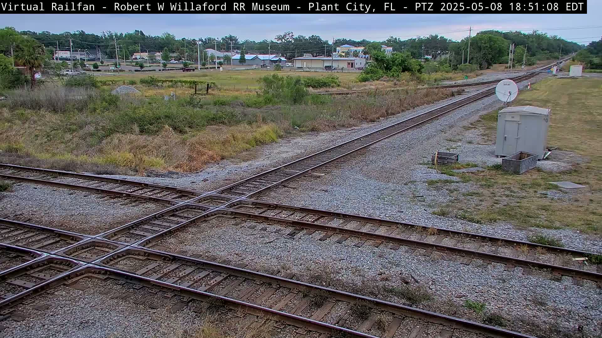 Train tracks intersect at a crossing, with a small building and satellite dish visible nearby under an overcast sky.