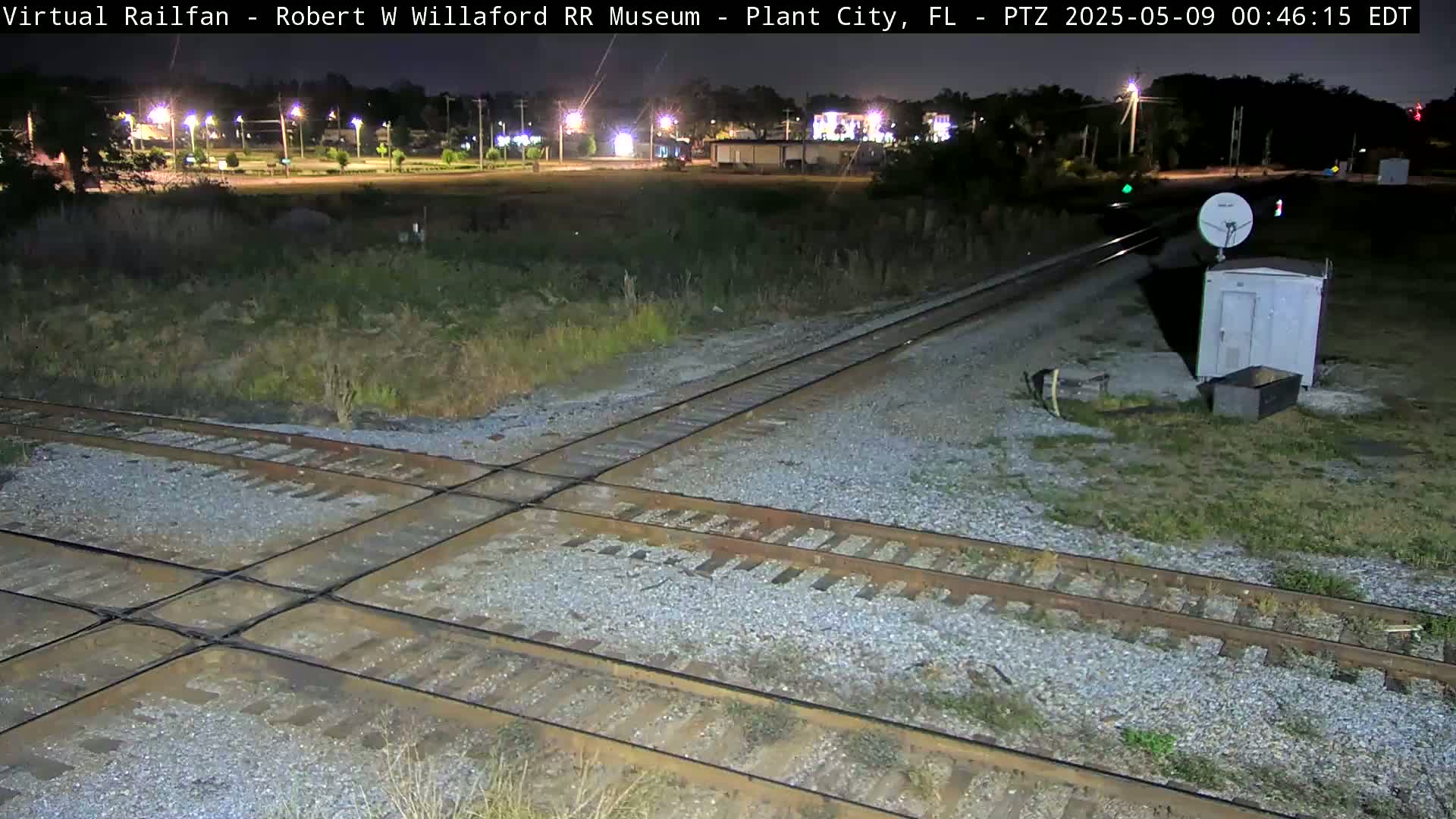 A nighttime view of a railroad crossing with a small building and satellite dish nearby, under clear skies.