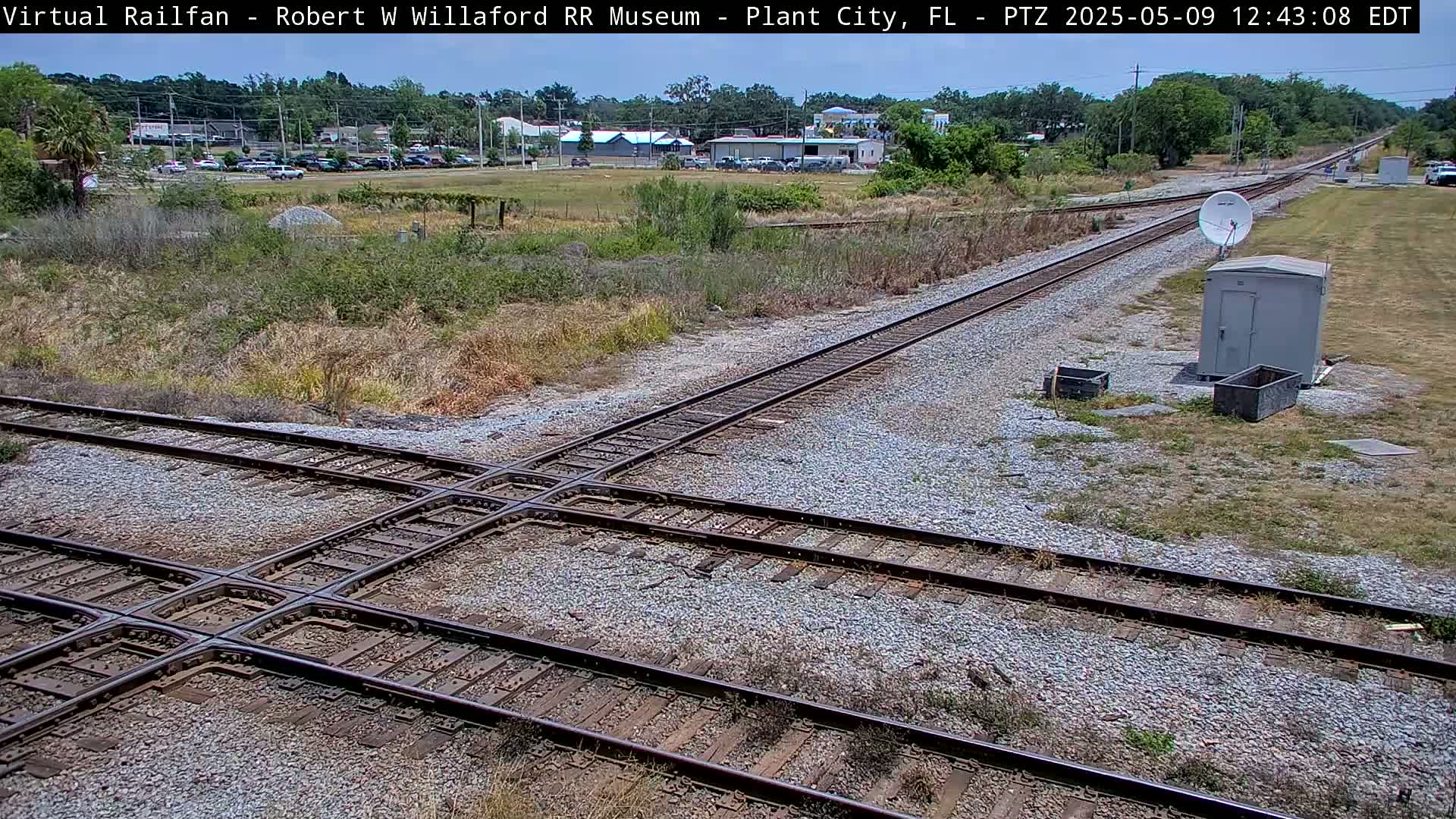 A railroad crossing intersects with two other sets of tracks under a sunny sky, surrounded by scrub brush and a small building with a satellite dish.