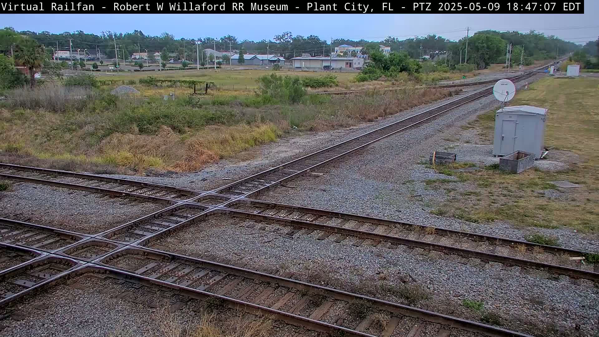 A railroad crossing with several tracks intersects amidst scrubby, dry vegetation under a partly cloudy sky.