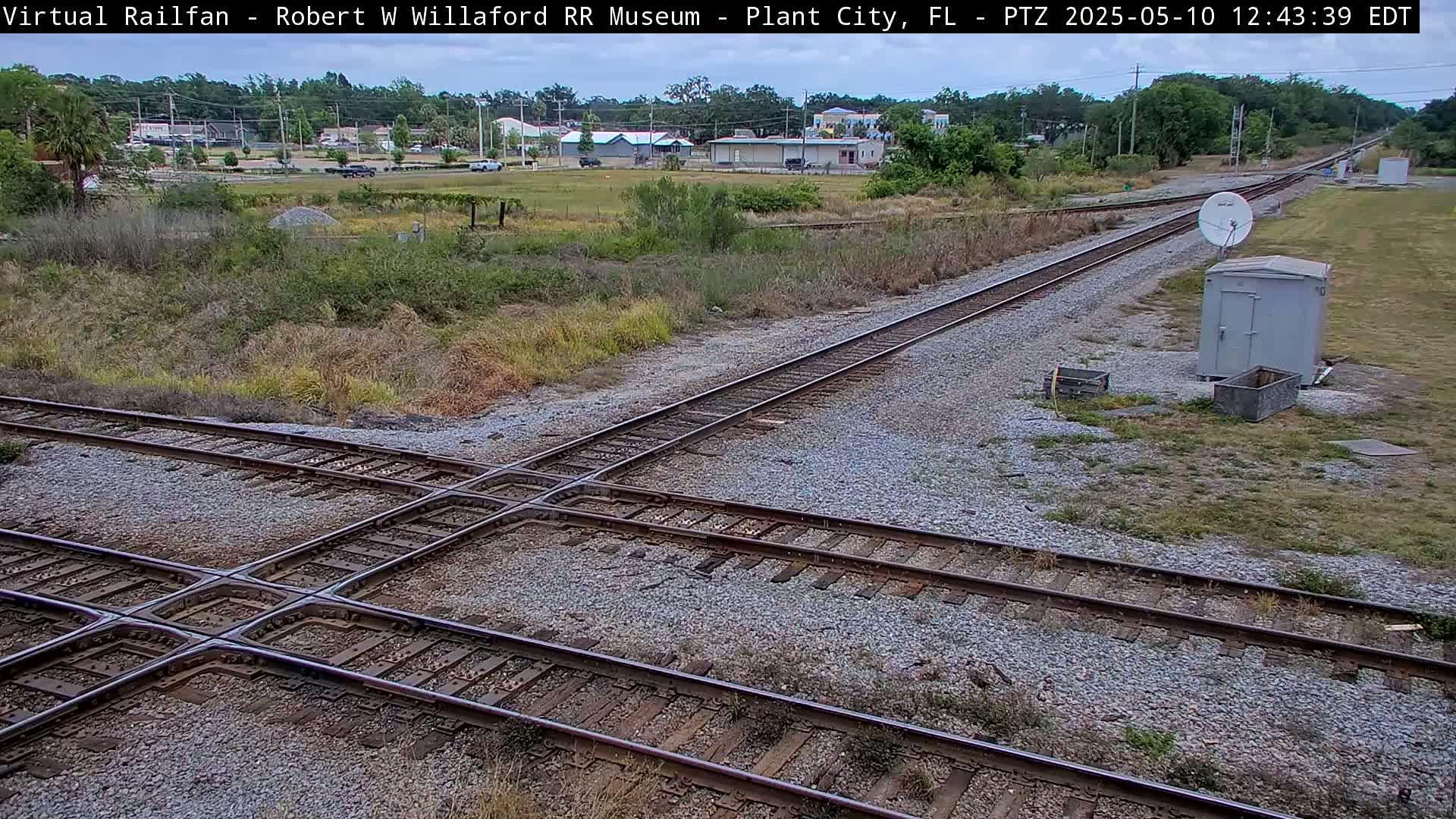 A high-angle view of a railway crossing with several tracks intersecting, surrounded by scrubby vegetation and a small building, under a partly cloudy sky.